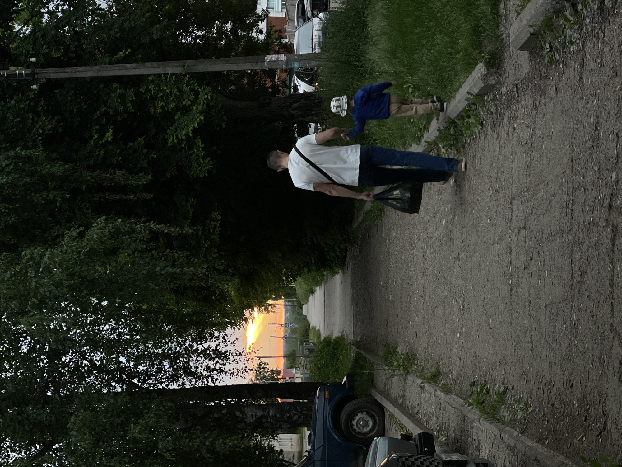This is a vertical, medium-distance, full-shot photograph taken at dusk in Chaikovsky, Russia, likely in a residential area. In the foreground, a man and a young child are walking away from the camera on a gravel path that runs alongside a concrete sidewalk. The man, wearing a white t-shirt and blue jeans, carries a black plastic bag in his left hand and holds the child's hand with his right. The child, facing away from the camera, wears a blue hooded jacket and tan pants, and a patterned bucket hat. They are walking towards a sunset visible in the distance, casting a warm orange glow.
To the left of the path, parked vehicles are visible, including a dark blue off-road vehicle. Trees with dense green foliage line both sides of the path, with a utility pole visible on the right side, partially obscured by leaves. The environment appears to be a street or pathway bordered by vegetation and residential buildings, suggested by the buildings visible in the far distance. The overall mood of the image is peaceful, with the soft light of the setting sun creating a serene atmosphere.