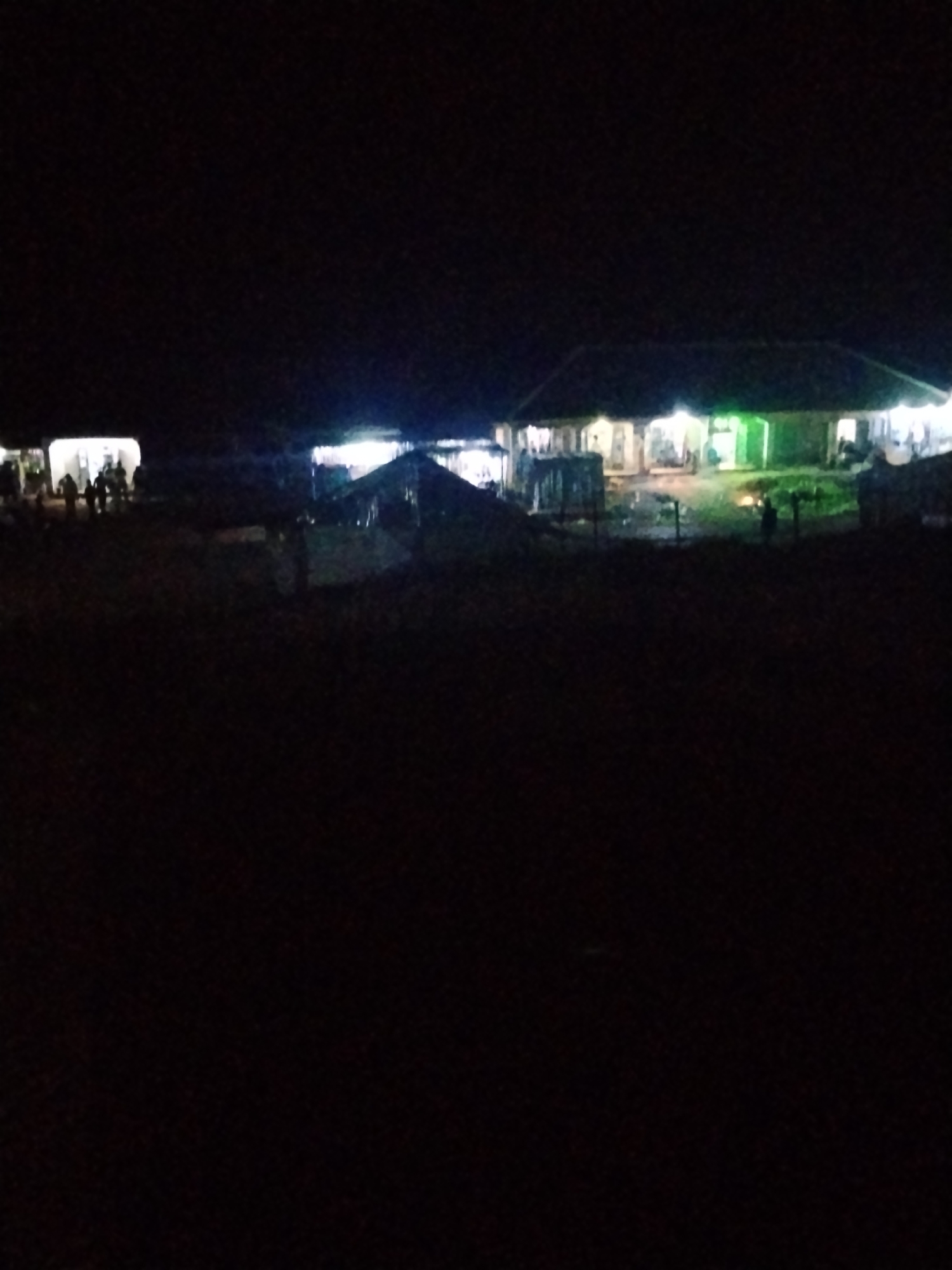 The image, taken at night, shows an outdoor gathering in what appears to be the city of Durumi, Nigeria. In the background, a building with a dark roof and illuminated windows suggests it is a place of business or residence. The light from the building casts a glow on the surroundings. Several people, appearing as silhouettes due to the darkness, are gathered in front of and around the building, indicating a social event or a market. Some structures, possibly stalls or temporary shelters, are also visible, illuminated by lights, suggesting some form of commerce or activity is taking place. The ground is dark, indicating it is likely unpaved or covered in dirt. The overall scene depicts a lively nighttime gathering in an outdoor, possibly semi-urban environment.