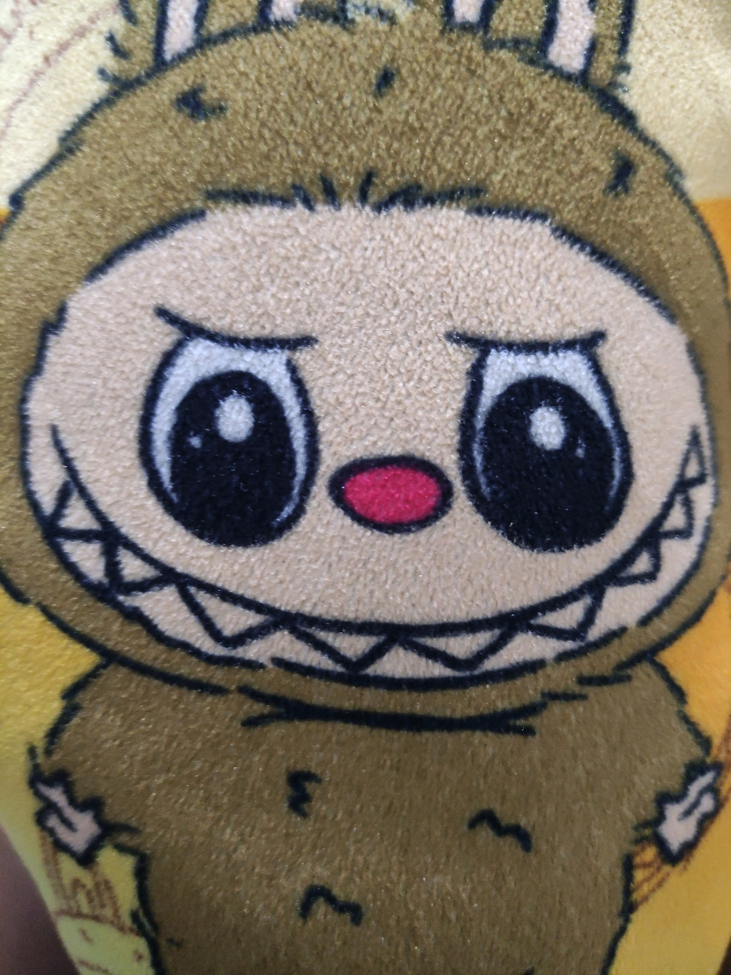 A close-up, full-frame, eye-level, medium shot captures a stuffed toy, possibly a pillow or plush. The toy depicts a creature with large, round black eyes outlined in white, a small red oval nose, and a wide mouth filled with sharp, triangular teeth. The creature is primarily brown and tan, with dark brown accents forming the outline of its face and features. The background is a bright yellow, and the overall impression is of a cartoonish, somewhat intimidating character. The image is sharp and well-lit, with a shallow depth of field, focusing on the texture of the plush material and the details of the drawn face. There is no discernible location within Bulukumba, Indonesia, or any specific environmental context.