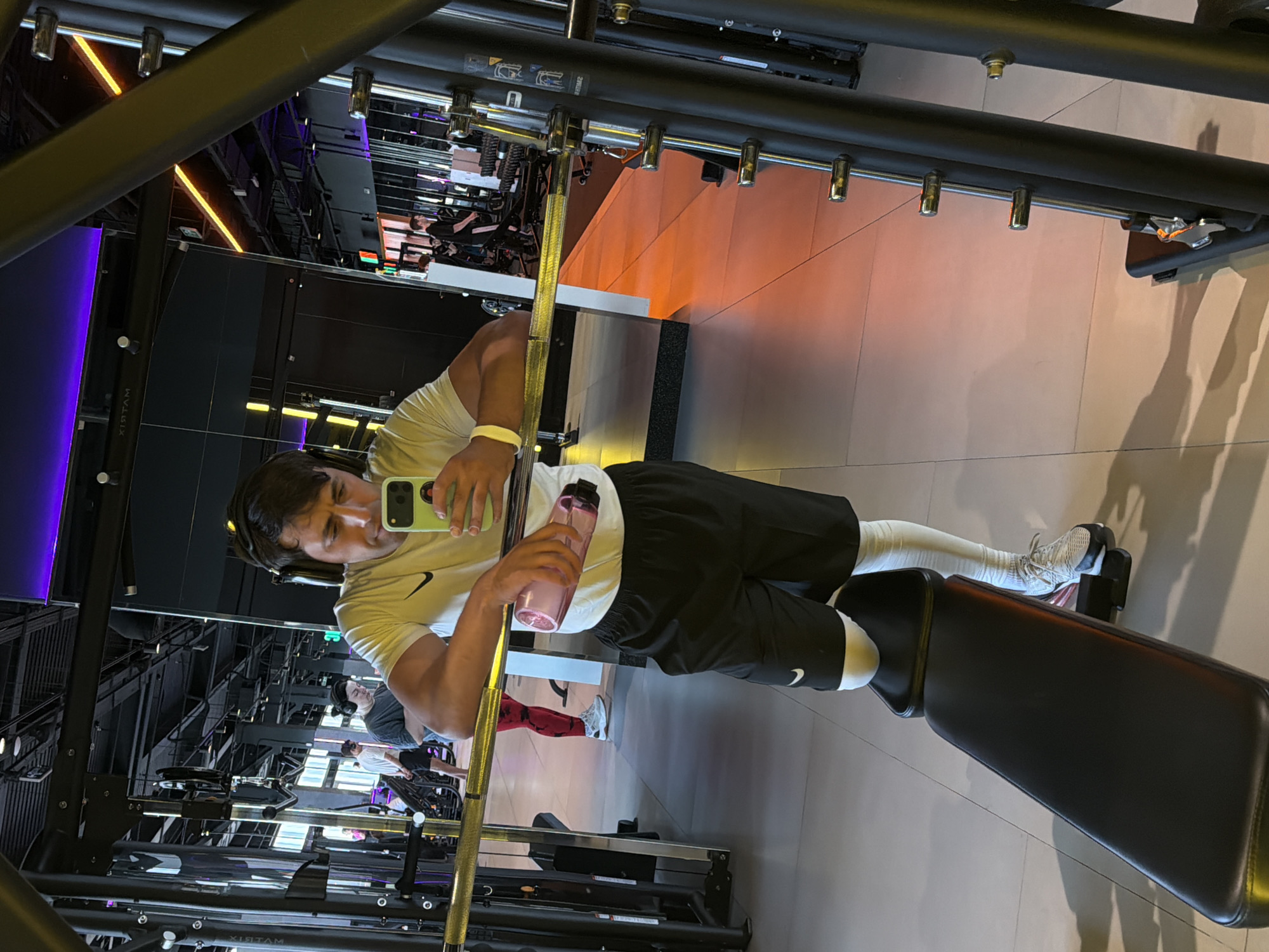 The image displays a selfie taken by a man in a gym mirror, captured in Krasnoyarsk, Russia. The man, appearing in the foreground, is shown from the waist up, dressed in a white t-shirt with a black Nike logo, black shorts with a white Nike swoosh on the left leg, white socks, and white sneakers. He has dark hair and headphones around his neck. In his left hand, he holds a light green smartphone, angled to take the picture, and in his right hand, he holds a translucent pink water bottle. A white wristband is visible on his right wrist.
The gym environment, visible through the mirror's reflection, is modern and well-lit. Dark walls and reflective surfaces create a sleek aesthetic. Various gym equipment, including racks and a black padded bench where the man's left leg rests, are scattered throughout. Other gym-goers are faintly visible in the background reflection, engaged in their workouts; one person is seen wearing red leggings. The lighting in the gym is a combination of white ambient light, vibrant purple strip lighting along walls and ceilings, and warm orange backlighting emanating from a distant wall section. The floor is a light, possibly tiled or polished concrete surface.
The activity taking place is a brief pause during a gym session, with the man capturing a moment with his phone. Visible text includes the Nike logo on his attire and the word "MATRIX" reflected on gym equipment. The word "SMUH" (likely reversed from an English word such as "HUMS" or part of an instruction) is also subtly visible on a machine in the background reflection.