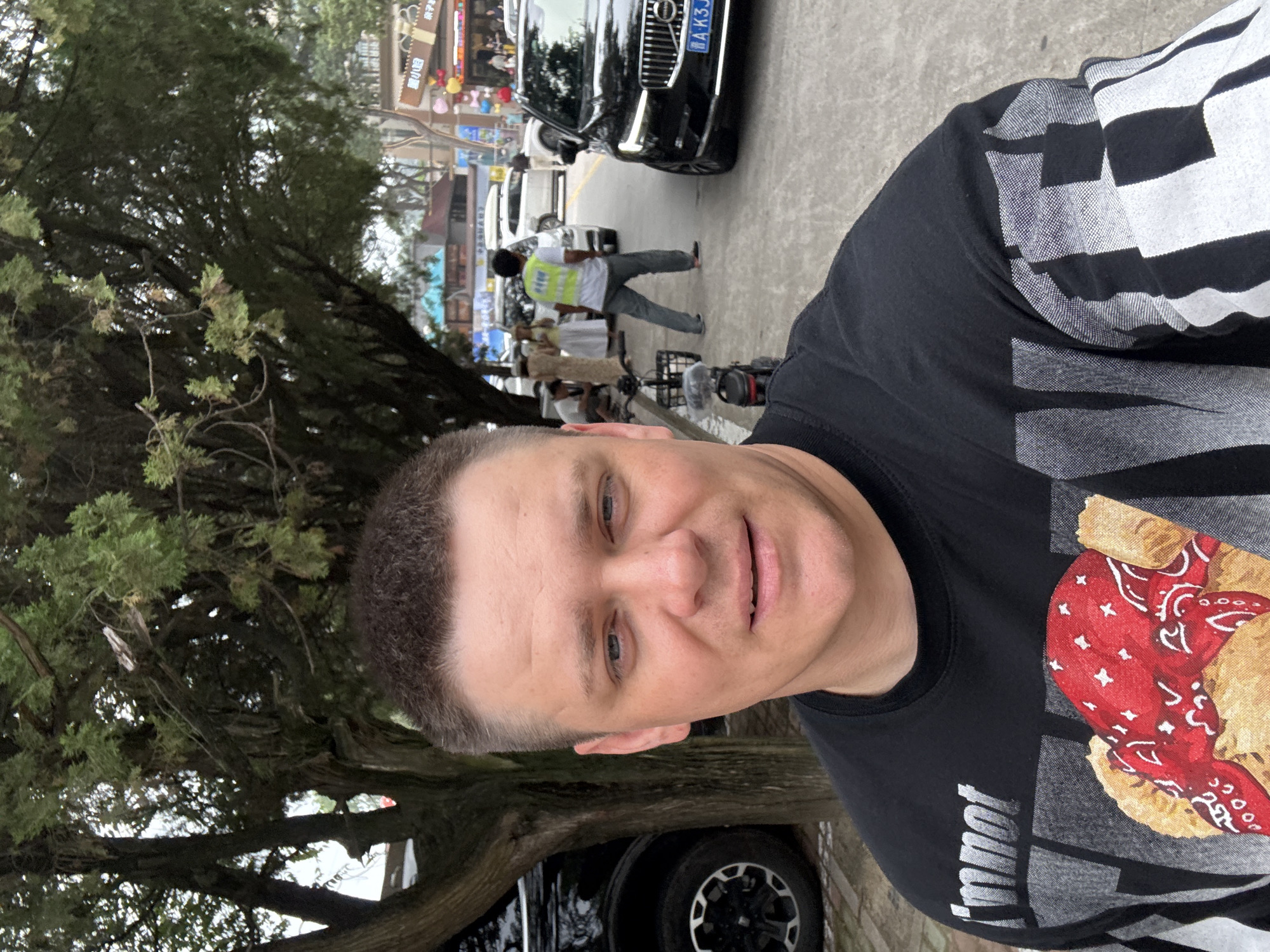 A man in a black t-shirt takes a selfie outdoors, with trees and a street scene in the background. The man has short brown hair and blue eyes. His t-shirt has a graphic design featuring text and an image of a teddy bear wearing a red bandana. In the background, there are several vehicles, including a black Volvo SUV with a license plate that reads "AK3JC". There are also people walking on the street and some shops visible. The scene appears to be in an urban environment. The location is identified as Qinhuangdao, China.