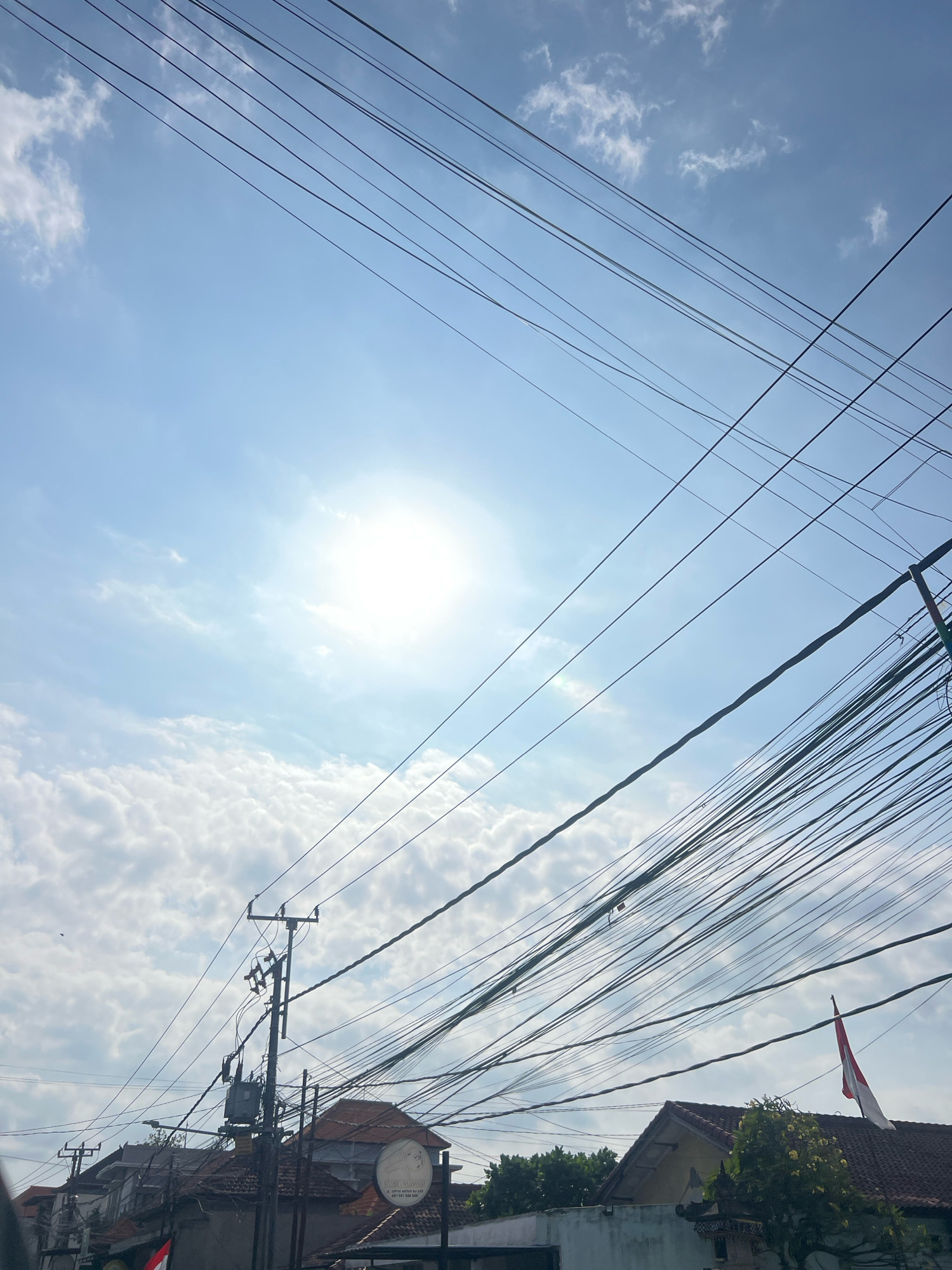 This image depicts a street scene in Denpasar, Indonesia, under a bright, sunny sky. Numerous utility wires, some appearing old and tangled, crisscross the frame, dominating the upper portion of the image. These wires are strung between utility poles, one of which is clearly visible with a transformer attached. In the background, the rooftops of residential buildings are visible, with some featuring tiled roofs. A small Indonesian flag is prominently displayed on one of the rooftops, waving in the breeze. The lower part of the image shows a building with a circular sign featuring text and an image. The overall impression is one of a densely populated urban area with a complex infrastructure of overhead wiring.