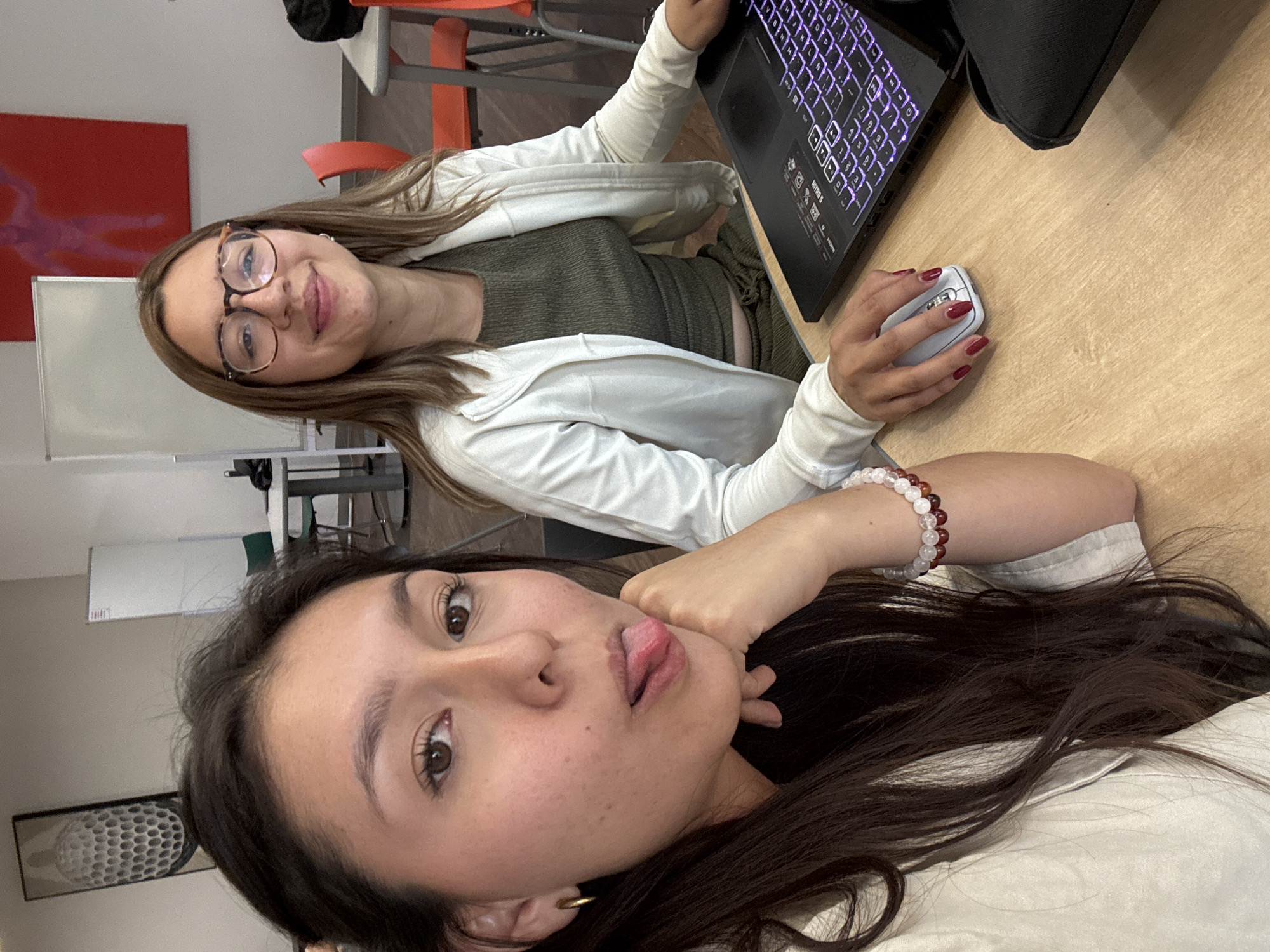 Two young women are depicted indoors at an educational or collaborative workspace in San Andrés Cholula, Mexico. The foreground features a woman with dark hair and a light-colored top, looking towards the camera with her tongue extended playfully, her right hand resting on her jawline. She wears a multi-colored beaded bracelet on her right wrist and a gold hoop earring. Seated to her right, a second woman with lighter brown hair and tortoise-shell glasses smiles at the camera while operating a black laptop with a glowing purple backlit keyboard and a white optical mouse with her left hand. She wears a white hooded jacket over a ribbed olive-green crop top and matching bottoms, and has dark red nail polish. The immediate environment includes a light-colored wooden table. The background reveals an off-white wall adorned with two distinct artworks: a framed, textured gray relief resembling a honeycomb or spherical pattern on the left, and a large red panel featuring a faint lighter-red human silhouette on the right. Additional elements include several tables with orange and green chairs, and a vertical whiteboard.