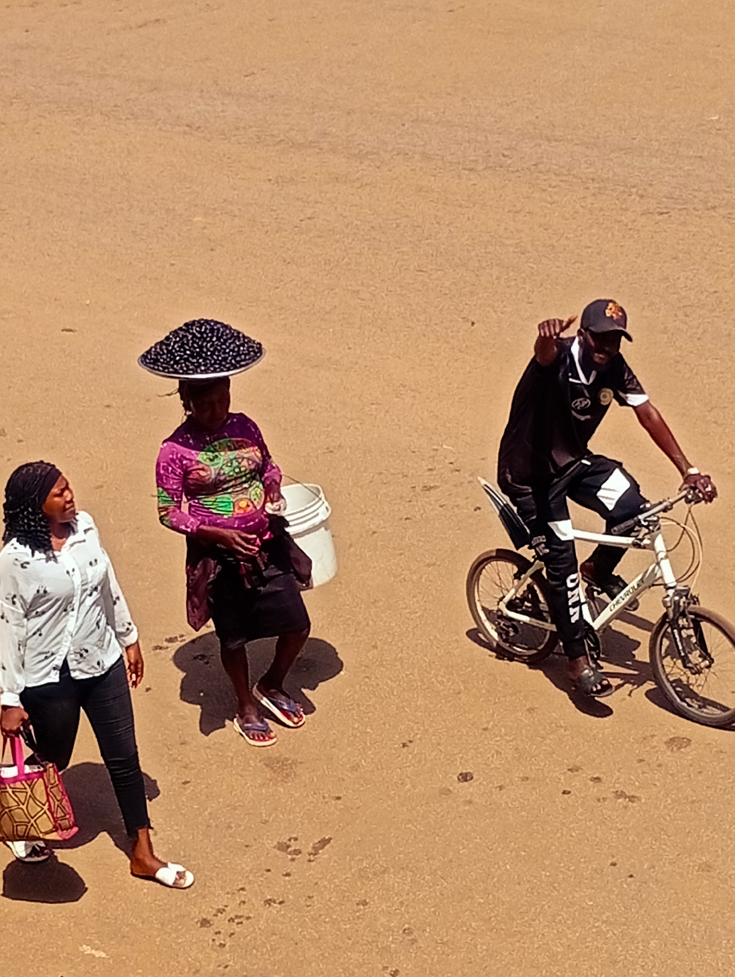 In Bukuru, Nigeria, an overhead shot captures a street scene with three individuals. The central figure, a woman, balances a large, circular tray overflowing with dark, round objects, likely berries or beans, on her head. She wears a vibrant, long-sleeved purple and green patterned tunic and a dark skirt. A white bucket hangs from her right arm. To her left, a woman in a white, collared blouse adorned with small black designs and dark pants walks towards the right, carrying a textured brown and pink bag in her left hand. To the right, a man on a white bicycle navigates the sandy ground. He wears a black baseball cap with an emblem, a black t-shirt with white accents, and black pants with white stripes. His left hand is raised, pointing forward. The ground is a dusty, reddish-brown, suggesting a dry, possibly sunny environment. The presence of multiple people suggests a typical day in a busy area, possibly a marketplace or a well-trafficked road. The bright sunlight casts distinct shadows on the ground, indicating the time of day is likely late morning or early afternoon. The overall mood appears casual, with individuals engaged in their daily routines. Visible text on the man's pants reads "DNA" and on the bicycle frame it reads "CHEVROLET".