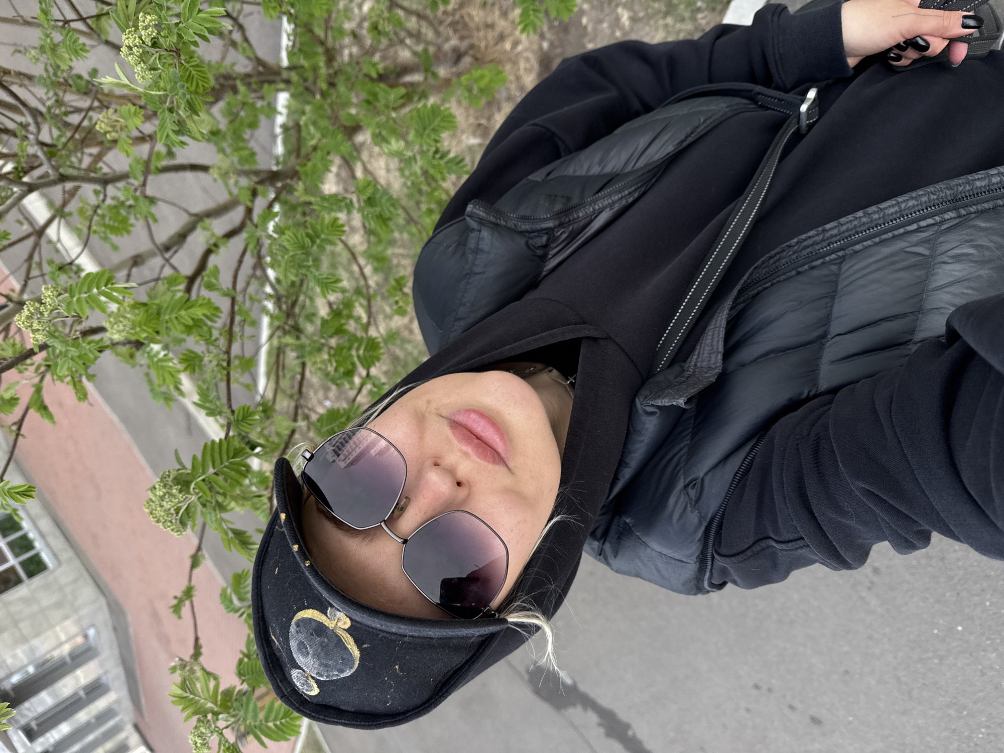A person with fair skin, wearing sunglasses and a black cap, is looking up at the camera. They are also wearing a black hooded sweatshirt and a black vest. They are standing outdoors, near a tree with green leaves. In the background is a pink building with white windows. The scene is likely located in a city in Krasnoyarsk, Russia.