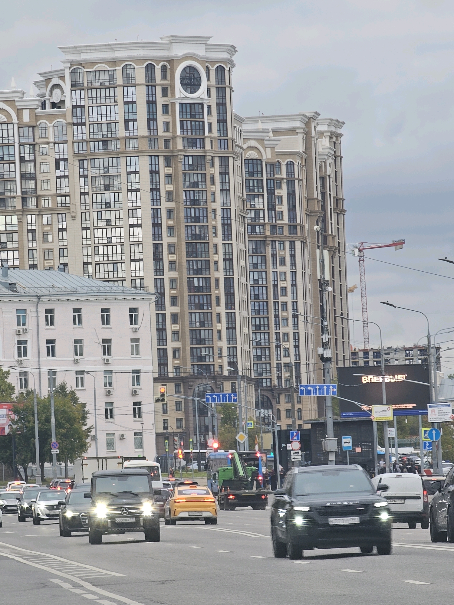 This is a street-level, eye-level medium shot of a busy, multi-lane road in Moscow, Russia. The scene is dominated by tall, modern buildings in the background, featuring a cream-colored facade with numerous windows and decorative architectural elements. A large construction crane is visible to the right of the buildings, suggesting ongoing development. The road is filled with various vehicles, including cars, trucks, and a yellow taxi. Headlights are illuminated on some of the vehicles. Traffic lights are visible, with one showing yellow. Road signs with directional arrows and traffic regulations are posted along the roadside. The sky is overcast with a neutral gray color. There are no visible people in the immediate foreground. The overall impression is a bustling urban environment.
