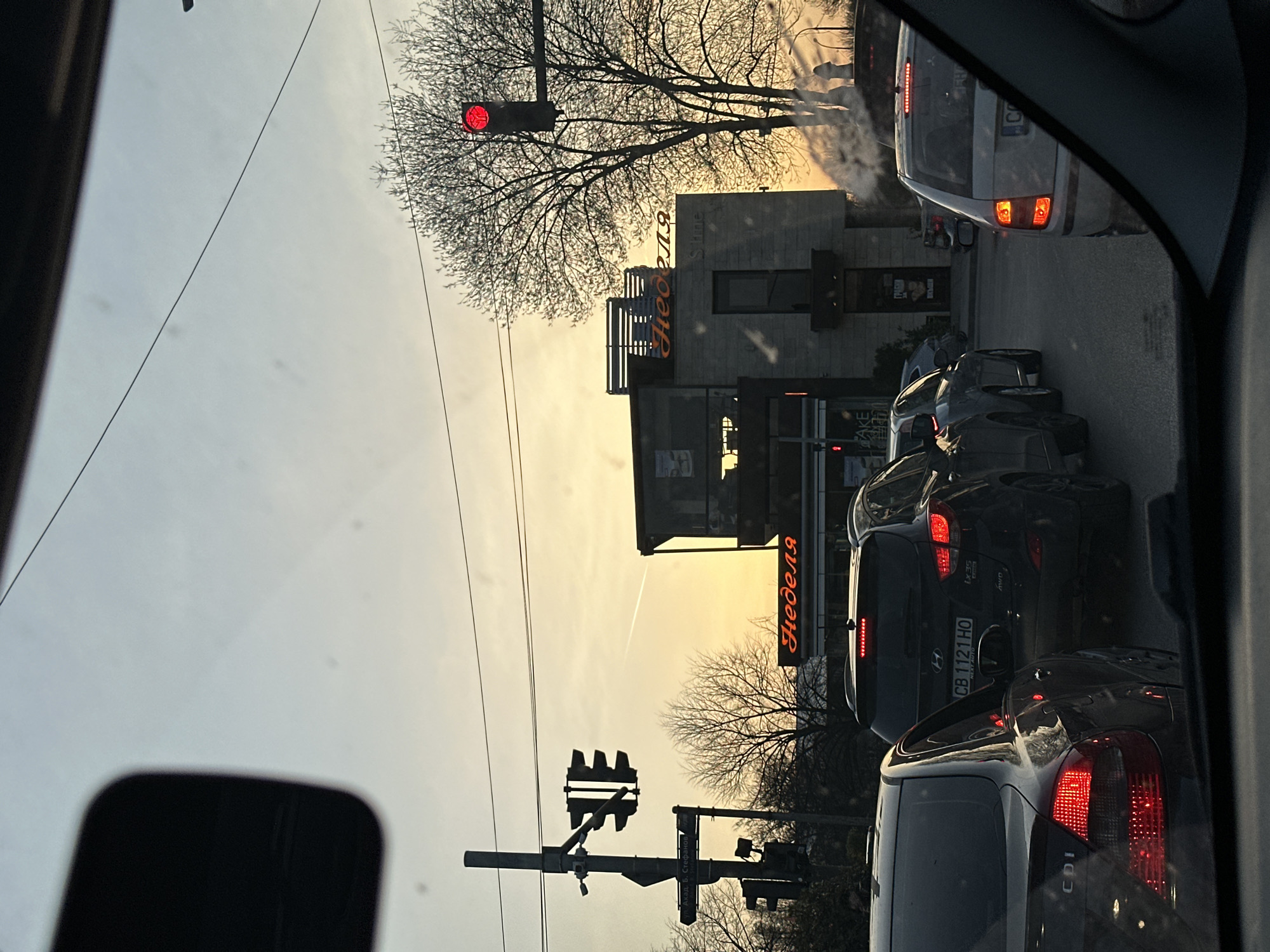 The image, taken from inside a car, shows a traffic scene in Sofia, Bulgaria. Cars are stopped at a red traffic light, under a sign written in Bulgarian Cyrillic: "Неделя" (Nedelya), identifying a confectionery chain. Another, smaller sign says "Stine." Visible license plates include "CB 1121 HO". Buildings are in the background, and a plane contrail is high in the sky. The light is pale from the rising or setting sun.