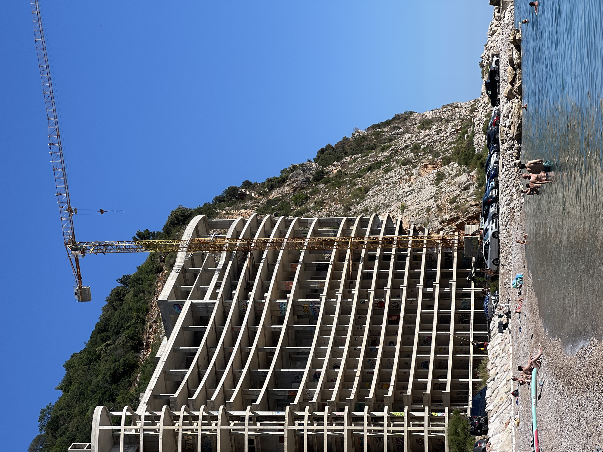 This image captures a multi-faceted scene on a clear, sunny day in Mirište, Montenegro, depicting a stark contrast between natural beauty, recreational activity, and incomplete infrastructure.
In the foreground, a pebbly beach meets calm, clear turquoise water. Several individuals are present: some are swimming or wading in the water, while others are seated on the beach. One person is sitting on a paddleboard. Beach towels and personal items are scattered on the pebbles.
Immediately behind the beach, a row of parked cars is visible on a rocky, elevated strip. Above the parking area, a massive, multi-story concrete building stands partially constructed, or possibly abandoned, directly at the base of a towering, rocky cliff. The building features a curved facade with numerous open levels, characterized by exposed concrete slabs and support columns. Many of the interior walls visible through the open structure are adorned with graffiti and colorful murals. A tall, yellow tower crane is positioned adjacent to the building, its long boom extending high into the clear blue sky, with a hook dangling.
The background is dominated by a very steep, rugged rock cliff face covered with scattered green trees and shrubs, rising sharply behind the construction and forming a natural boundary to the scene. The sky above is a uniform, cloudless deep blue. The overall impression is one of a tourist destination undergoing significant development, with recreational activities occurring simultaneously next to a large-scale construction or abandoned project against a dramatic natural backdrop.