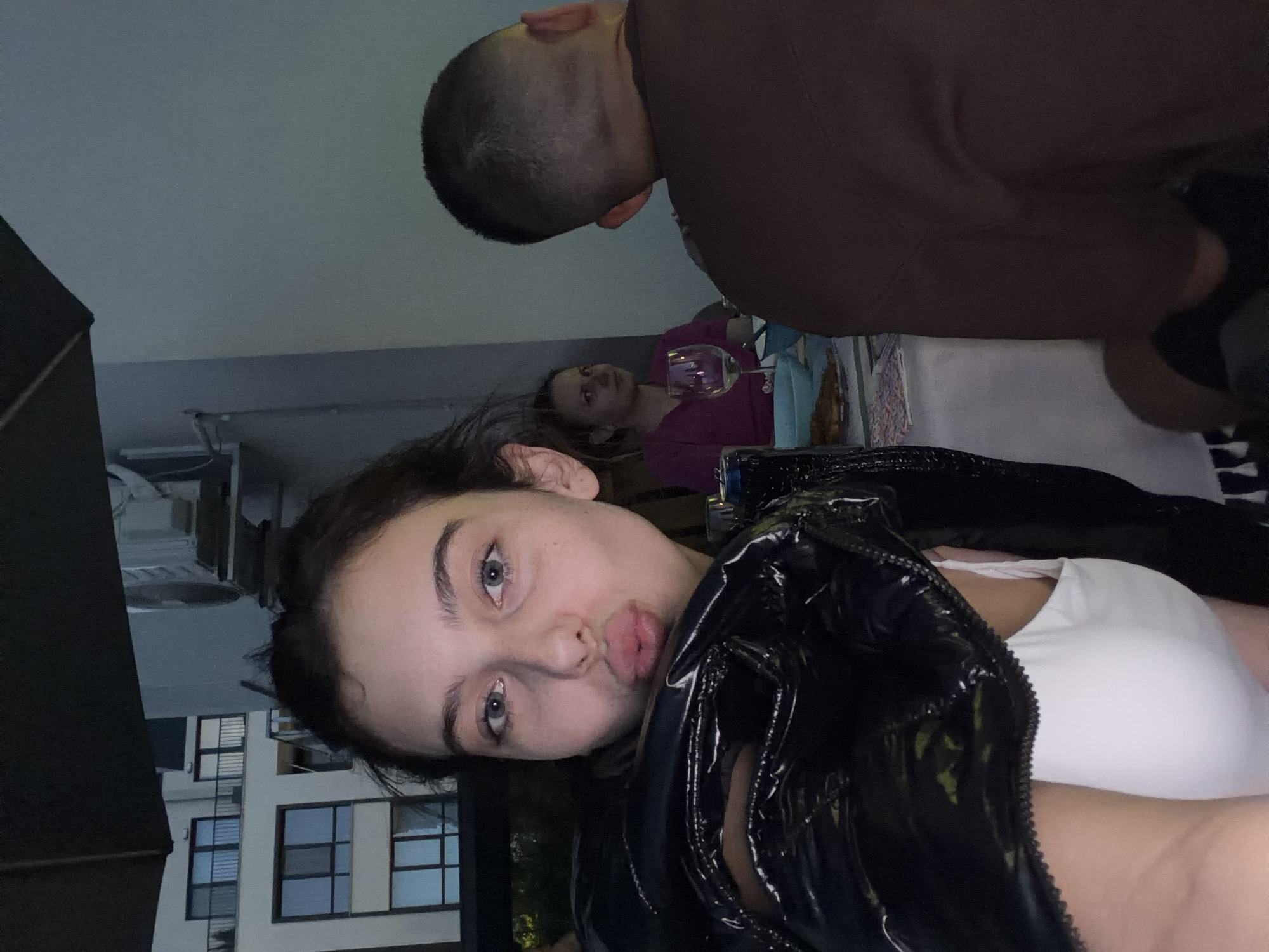 The media file is a photograph taken indoors, likely at a party or gathering. The foreground is dominated by a young woman with dark hair and blue eyes, who is looking at the camera with her lips pursed, as if blowing a kiss. She is wearing a white top and a shiny black jacket. To her left, a portion of a dark, possibly fabric object is visible. In the background, two other individuals are partially visible. One, a man with a shaved head, is leaning forward, and another person, a woman wearing a purple top, is holding a wine glass. The background also shows elements of a domestic setting, including what appears to be a table and possibly part of a building through a window. The lighting suggests it is evening or nighttime. The overall mood appears casual and social. No text is visible in the image.