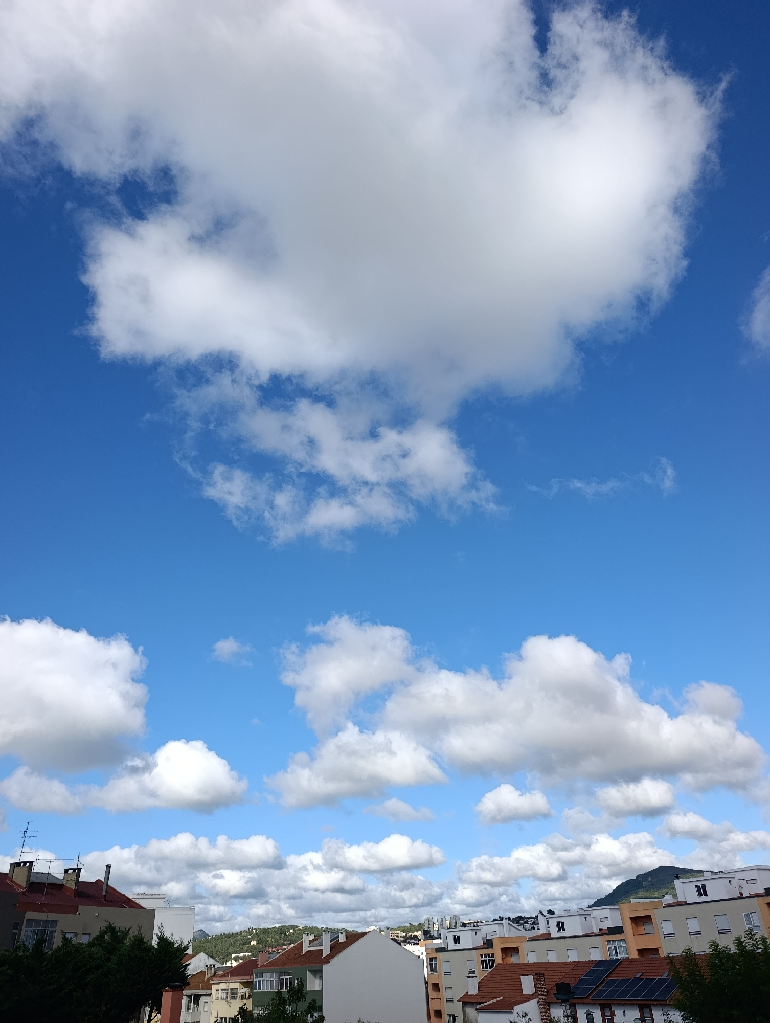 This image captures a view of rooftops and a blue sky filled with fluffy white clouds in Setúbal, Portugal. The buildings are a mix of architectural styles, with some featuring red tile roofs and others with flat white roofs. Solar panels are visible on one of the rooftops. The sky is a vibrant blue, dotted with numerous clouds of varying sizes and densities, some appearing bright and sunlit while others are shadowed. In the background, a gently sloping hill covered in trees can be seen, adding a touch of natural landscape to the urban scene. The perspective is from a slightly elevated viewpoint, looking over the tops of the buildings towards the sky and distant hills.