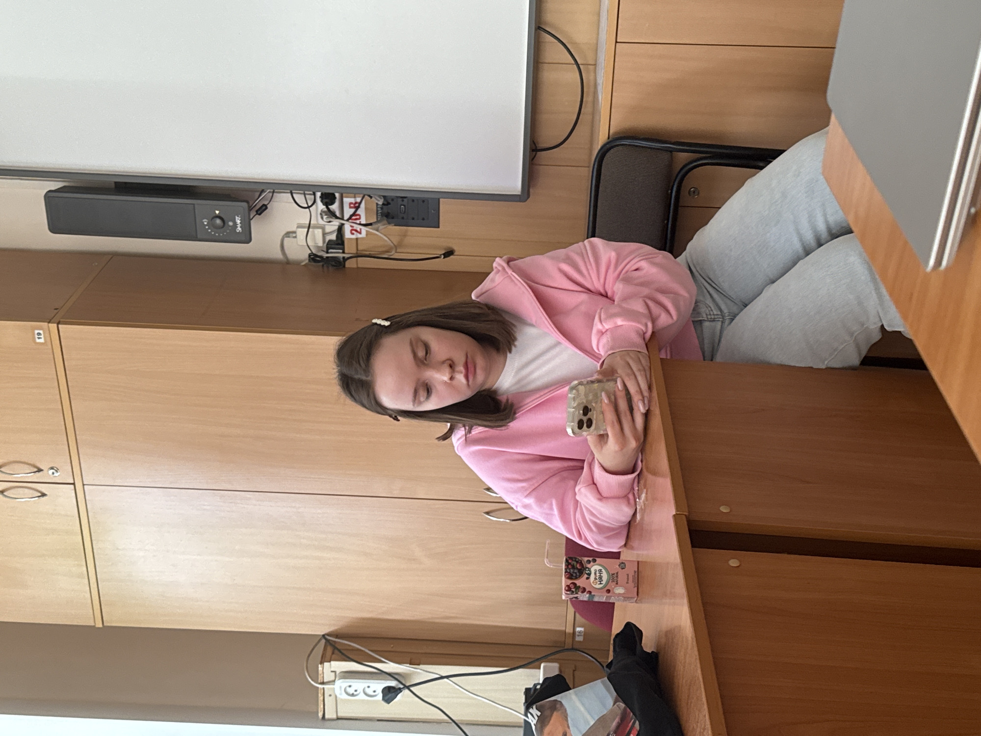The image, rotated 90 degrees counter-clockwise, captures a young woman resting at a wooden desk in what appears to be a classroom or study area. She has brown hair, adorned with a small pearl-like clip, and is dressed in a pink hooded jacket over a white top and light blue jeans. Her eyes are closed, suggesting she is sleeping or deeply resting, and her hands are clasped around a smartphone encased in a clear, possibly glittery, cover.
The setting includes a white board mounted above a black rectangular soundbar or speaker labeled "SMART," which has a visible knob. Below the soundbar, multiple electrical outlets, power strips, and tangled wires are visible against the wooden wall. A red label with "24В" is affixed near the outlets. A silver-colored laptop is partially visible on the right side of the desk.
On the desk in front of the woman, there is a small carton of berry-flavored juice or beverage. The carton displays Russian text "ЛЮБИМЫЙ НАППИ," along with "100% NATURAL," "100% НАТУРАЛЬНЫЙ," and "ФРУКТОВЫЙ." Wooden cabinets with silver handles are visible to the left. A black chair with a mesh back is positioned behind the woman. Partially obscured beneath the desk are a black bag or fabric and a piece of paper. The indoor environment provides no specific cues for time of day or weather, but the classroom setup and the woman's relaxed, sleepy demeanor are central to the scene. The visible Russian text on the beverage carton grounds the location context to Perm, Russia.