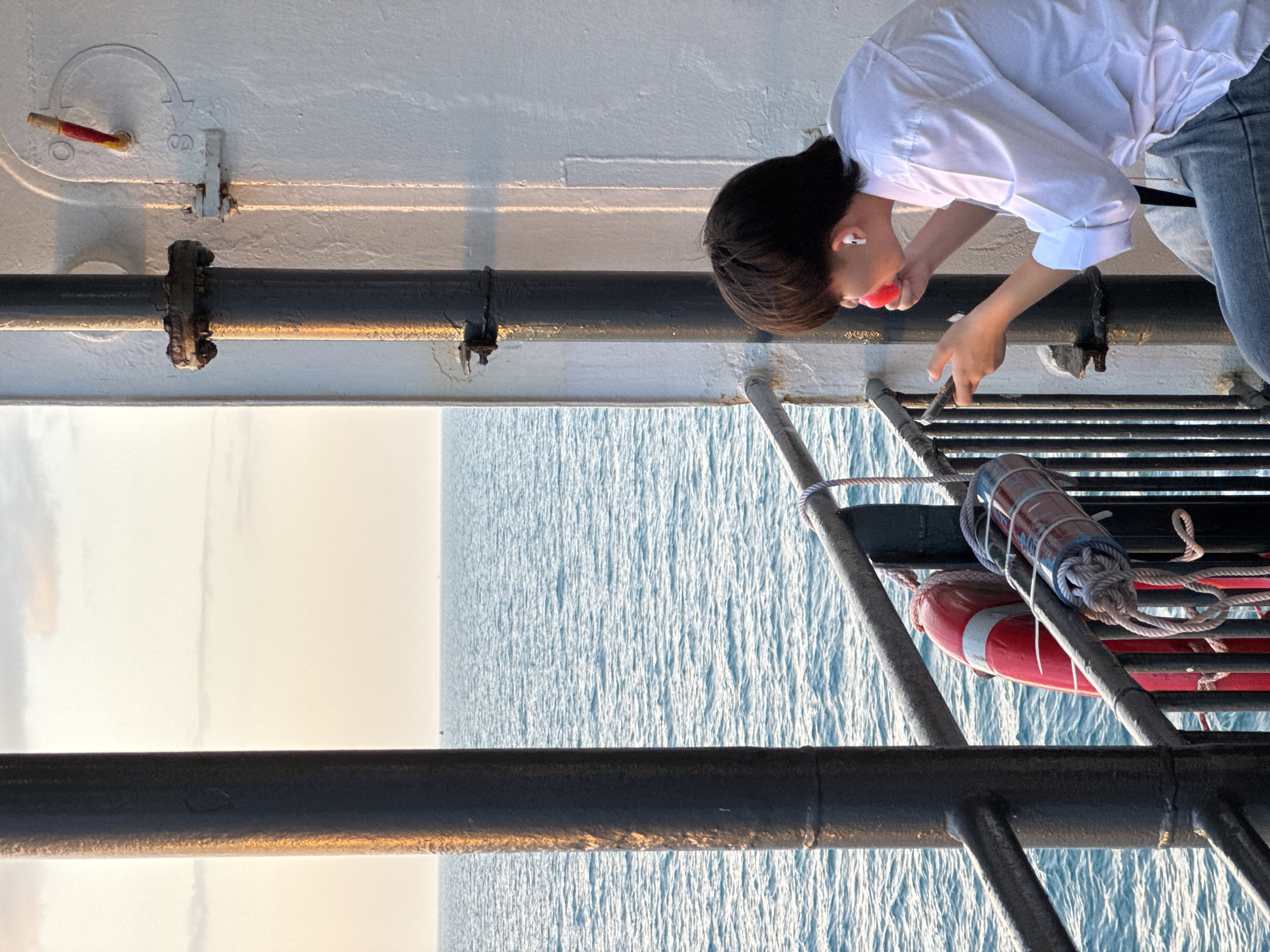 A young person with short, dark hair, wearing a white collared shirt and denim jeans, is seated on a ferry deck. They have white earbuds in their ears and are holding a red spherical object to their mouth with their right hand, while looking at a smartphone in their left hand. The ferry is sailing on a blue ocean under a pale orange and white sky. A black metal railing runs along the deck, and a red life preserver with a white band and a cylindrical life-saving device are attached to it. The ferry's white outer wall is visible on the right, with a vertical pipe and some attached hardware. The scene is illuminated by the warm light of a sunset or sunrise.