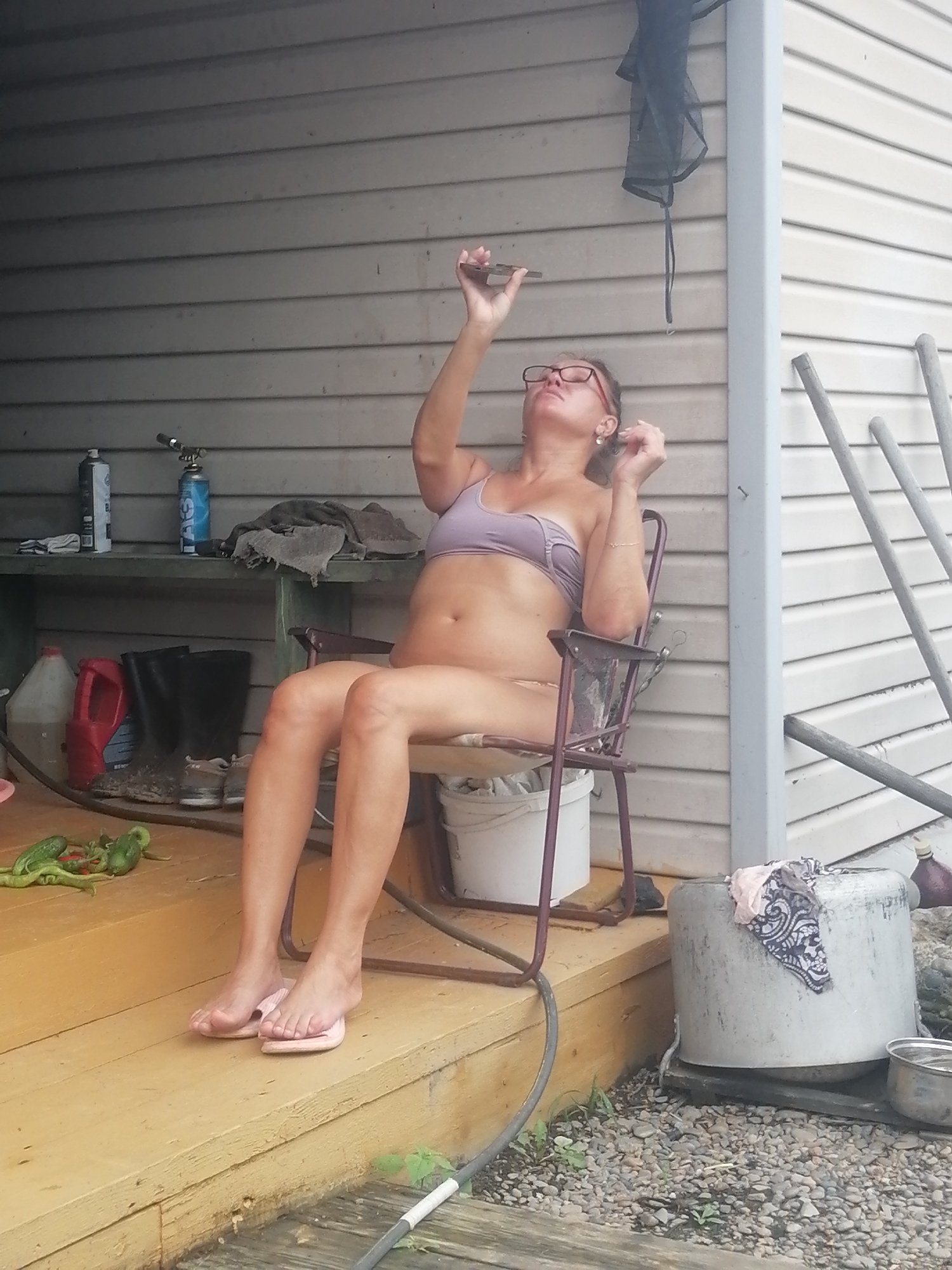 This media file depicts a woman sitting on a chair on a wooden porch. She is wearing glasses and a light purple bikini top, and her lower body is exposed. Her right arm is raised, and she appears to be holding a phone or small object. The setting is outdoors, with a light-colored, horizontal-paneled building behind her. To her left, on a shelf, are two spray cans, one blue and one white, and some dark fabric. Below the shelf, there are several containers, including red and clear jugs, and a pair of black boots. In front of the porch, on the wooden planks, are some green vegetables and a black hose. To the right of the woman, a large metal container with a patterned cloth draped over it sits on the gravel ground. The overall lighting suggests it is daytime. There are no visible text elements.