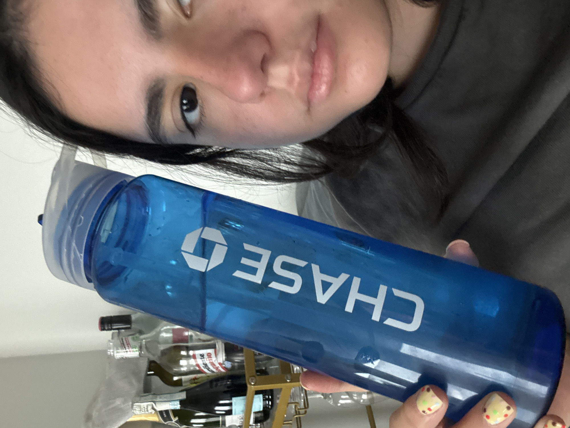 An eye-level, rotated shot captures a person holding a blue water bottle. The image is rotated roughly 90 degrees clockwise, with the person's face visible in the upper left, angled towards the viewer. The person has dark hair, dark eyes, and a neutral expression. They are wearing a dark grey or black t-shirt.
Their right hand is visible in the lower right, holding the blue, translucent water bottle that extends diagonally across the frame. The bottle has "CHASE" written in white capital letters on its side, along with the associated Chase bank logo. The bottle contains some liquid. The person's fingernails are painted light yellow/cream with small, colorful designs, possibly resembling fruit.
In the blurred background, behind the water bottle, there is a gold-colored rack holding several glass bottles, which appear to be wine or liquor bottles. Some faint, blurry text is visible on the labels of these background bottles, including fragments like "diente" and "guetto" or similar. The setting appears to be indoors, possibly a domestic environment or a casual space where beverages are stored.