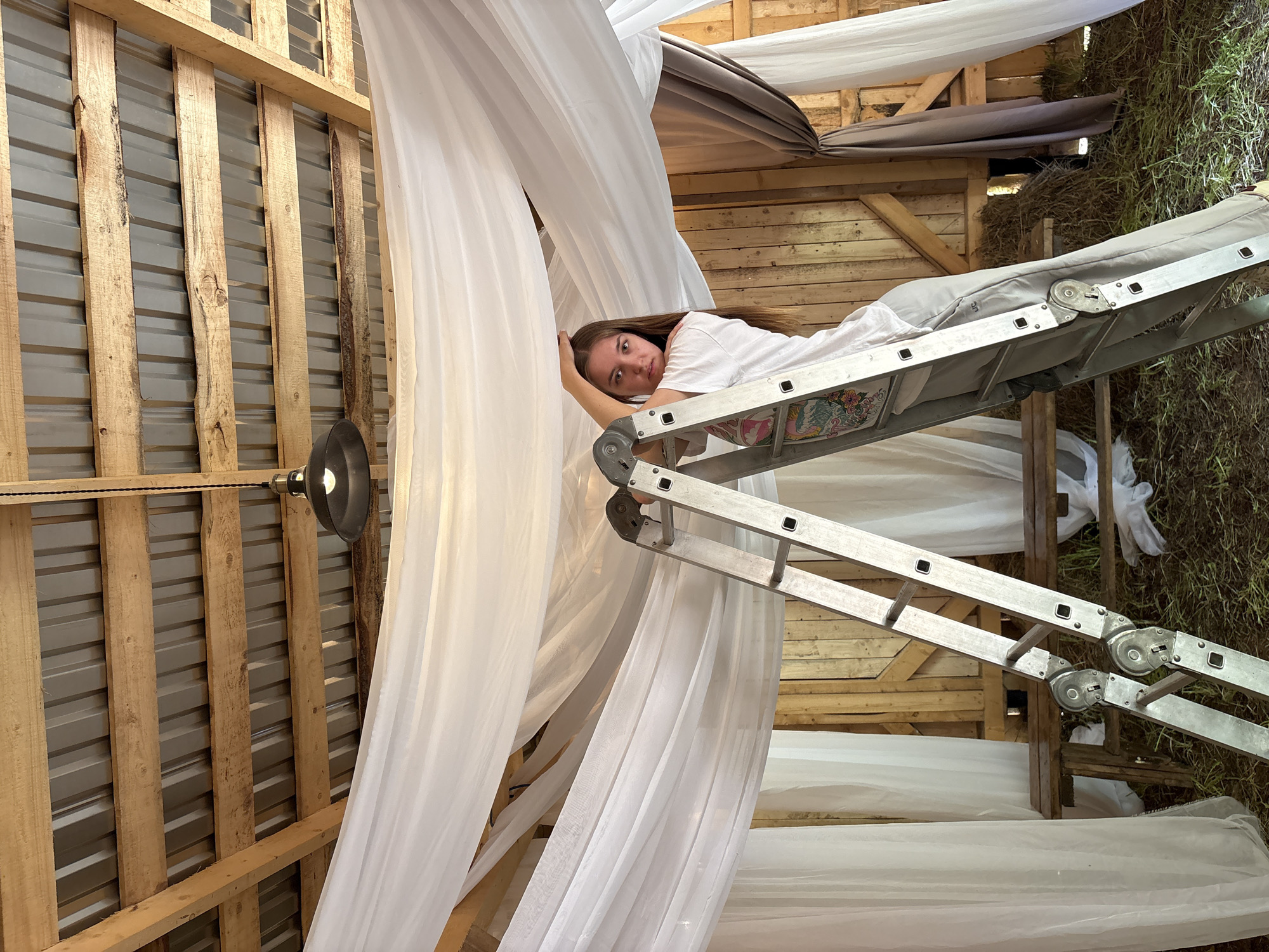 A young woman lies on a ladder within a wooden structure in Sibir', Russia. The ladder is angled, resting against the structure's walls and ceiling. The space is adorned with draped white fabric, creating a soft, ethereal atmosphere. Hay is visible on the right side of the frame. A black lamp fixture hangs from the ceiling on the left. The woman is wearing a white shirt. The walls of the wooden structure are constructed of a combination of wood and corrugated metal sheeting.
