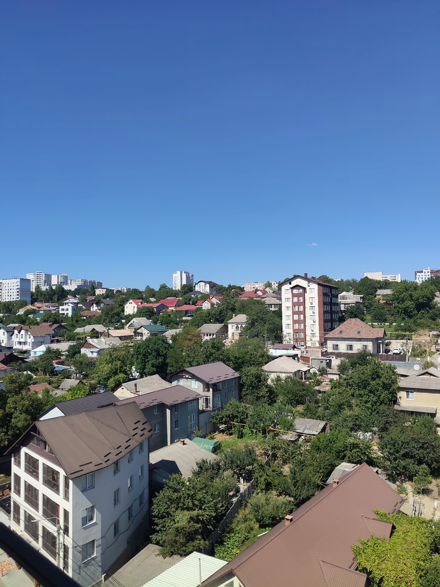 This is a high-angle, landscape photograph of a residential area with a clear blue sky. The scene depicts a densely packed neighborhood with a mix of buildings, including multi-story apartment buildings in the background and numerous houses with sloped roofs in the foreground and midground. The houses are of varying sizes and architectural styles, with many featuring visible windows and chimneys. Lush green trees are interspersed throughout the neighborhood, creating a verdant environment. The photograph appears to have been taken on a sunny day, with the sunlight casting shadows on the buildings and foliage. The image captures a typical urban residential landscape with abundant greenery.