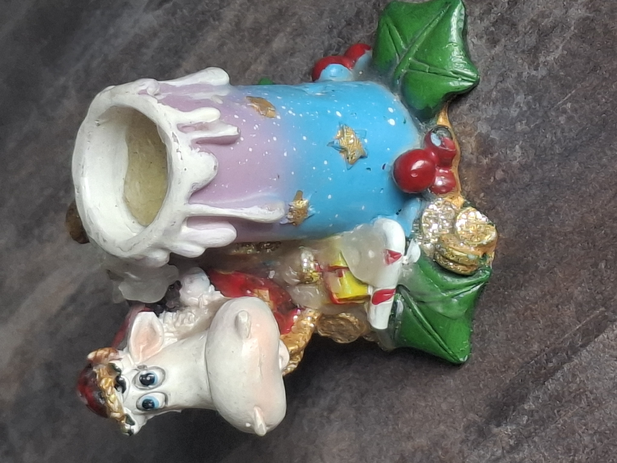 This is a close-up, eye-level photograph of a decorative candle holder. The candle holder is shaped like a white cow wearing a red and gold hat and a red garment. The cow is positioned to the left and is looking towards the viewer with blue eyes. In front of the cow are various small, colorful objects resembling gifts. To the right of the cow is the candle holder itself, which is cylindrical and decorated with a gradient of pink and blue colors, dotted with white specks and small gold stars. White wax drips down the sides of the candle holder. The base of the candle holder is adorned with green holly leaves and red berries. The object is placed on a dark, textured surface that appears to be wood or a similar material. There are no people or animals present in the image. The image does not contain any information that would suggest a location in Almaty, Kazakhstan.