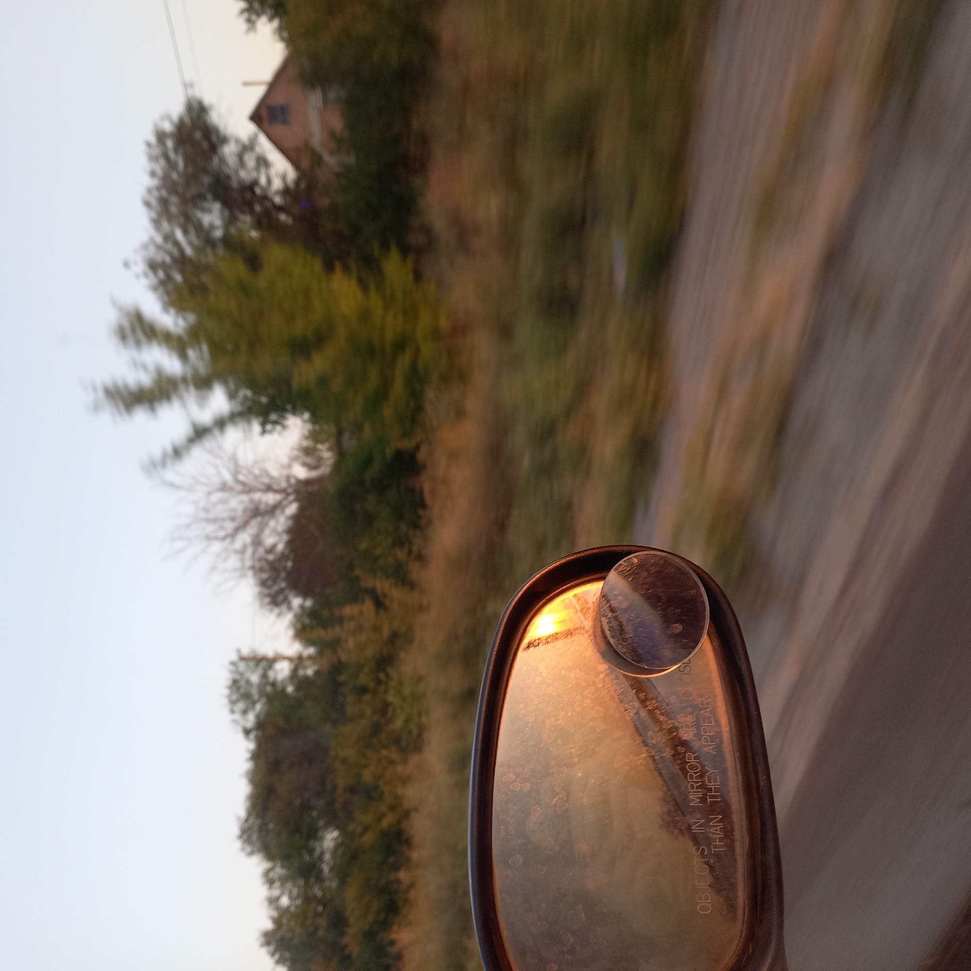 The image shows a close-up view of a car's side mirror, reflecting the setting sun and a blurred rural landscape. The mirror is dirty, with visible smudges and water spots. The text "OBJECTS IN MIRROR ARE CLOSER THAN THEY APPEAR" is inscribed on the mirror. In the reflection, a house with a peaked roof and a chimney is visible amidst trees, suggesting a rural or semi-rural setting. The background of the image is motion-blurred, indicating the car is in transit, likely on a dirt or gravel road given the texture of the foreground. The sky is a pale blue, transitioning to a warm orange hue from the setting sun. The overall impression is one of a journey through a rural area at dusk. The specific location in Sonyachne, Ukraine cannot be determined from the visual information alone.