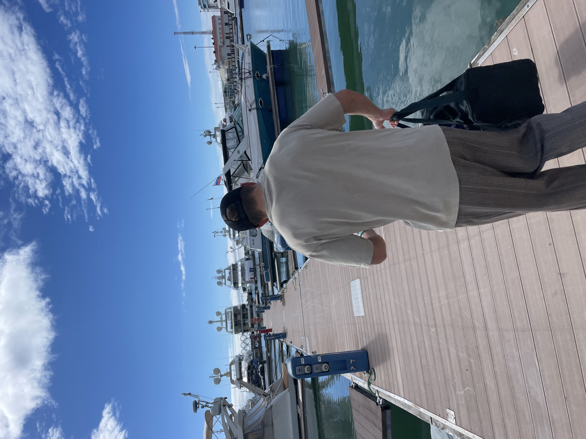 A man in a light beige short-sleeved shirt, dark pants, and a black baseball cap walks on a wooden dock. He carries a black duffel bag. The dock is lined with boats and yachts. In the background, there are buildings and a bright blue sky with scattered clouds. The location is Sochi, Russia.