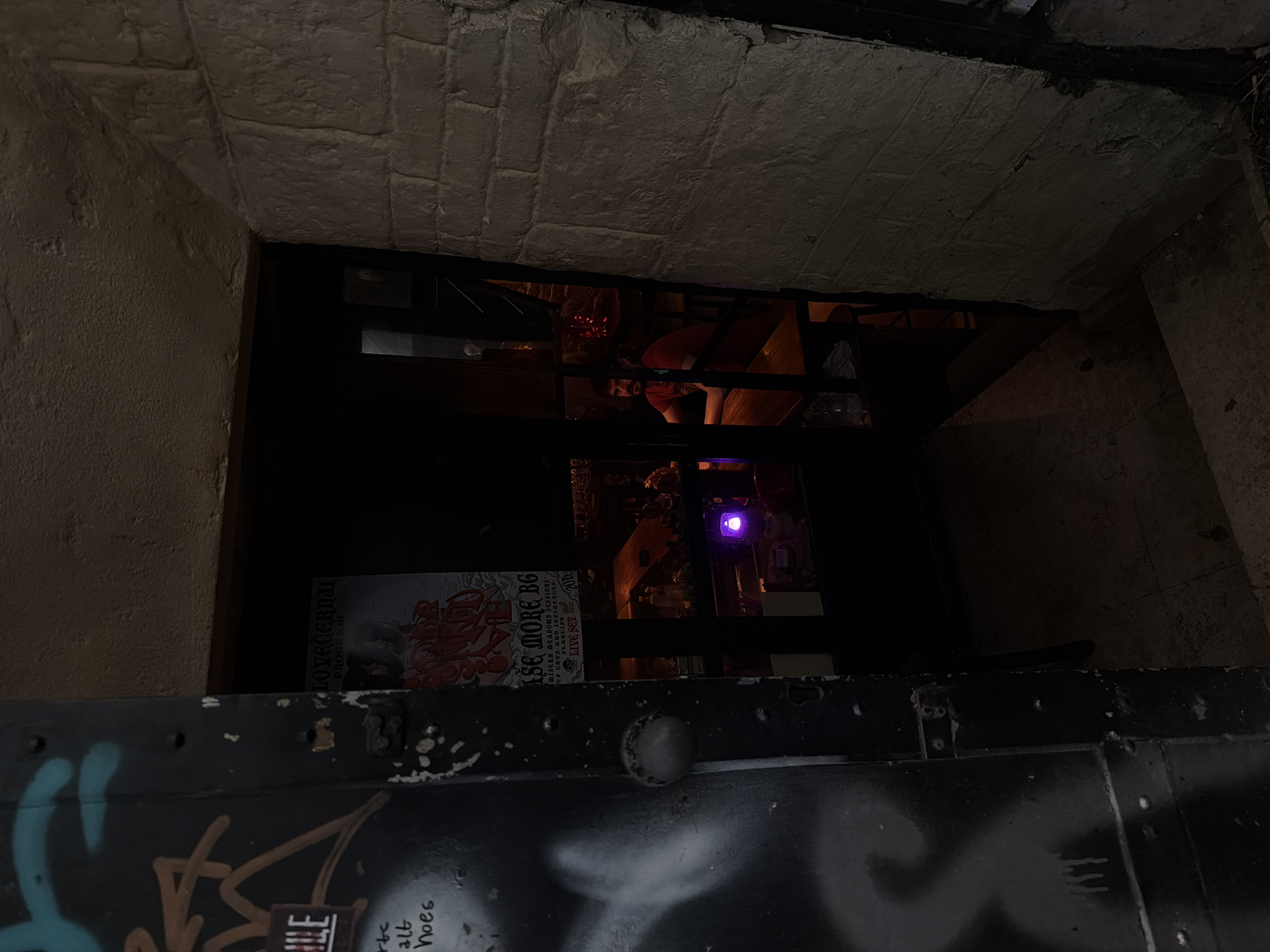 Captured from a low, street-level vantage point in Belgrade, Serbia, this dimly lit, urban scene reveals a glimpse inside a possible bar through a dark doorway. The perspective is partially obstructed by what appears to be a metal, graffiti-covered barrier or door featuring prominent rivets and a single, silver knob. Graffiti art, including partial words such as "ILE," "rtc," and "hoes" as well as "LOVEČERNJI PROGRAM" is visible on this barrier.
Through the dark doorway, the interior of a dimly lit space is visible. A red-shirted individual can be seen among the furniture and other people. A purple light fixture is also discernable in the dark interior. The overall environment suggests an intimate or hidden space, and the setting is seemingly in Belgrade, Serbia.