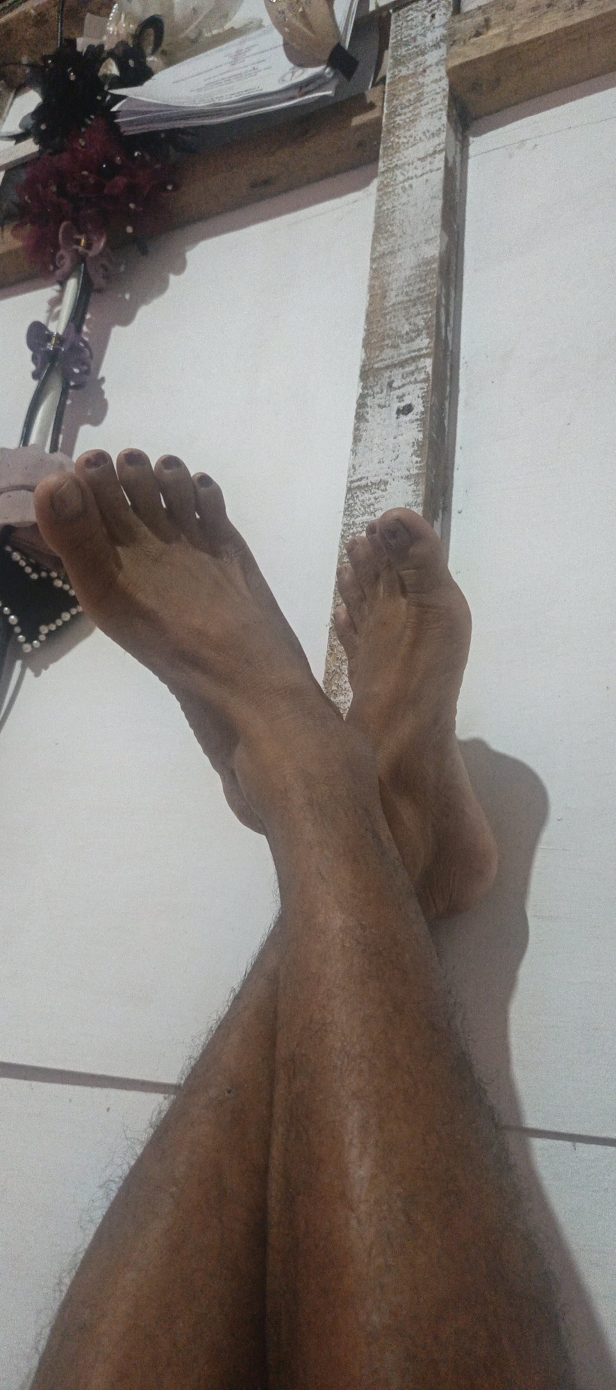 In Cinta Dame, Indonesia, a close-up shot focuses on a person's crossed feet and lower legs, positioned against a white wall. The feet are bare, with toenails appearing to have dark nail polish. The legs are covered in dark hair. The background includes a wooden structure and a stack of papers or documents. The lighting casts a shadow of the crossed legs on the wall.