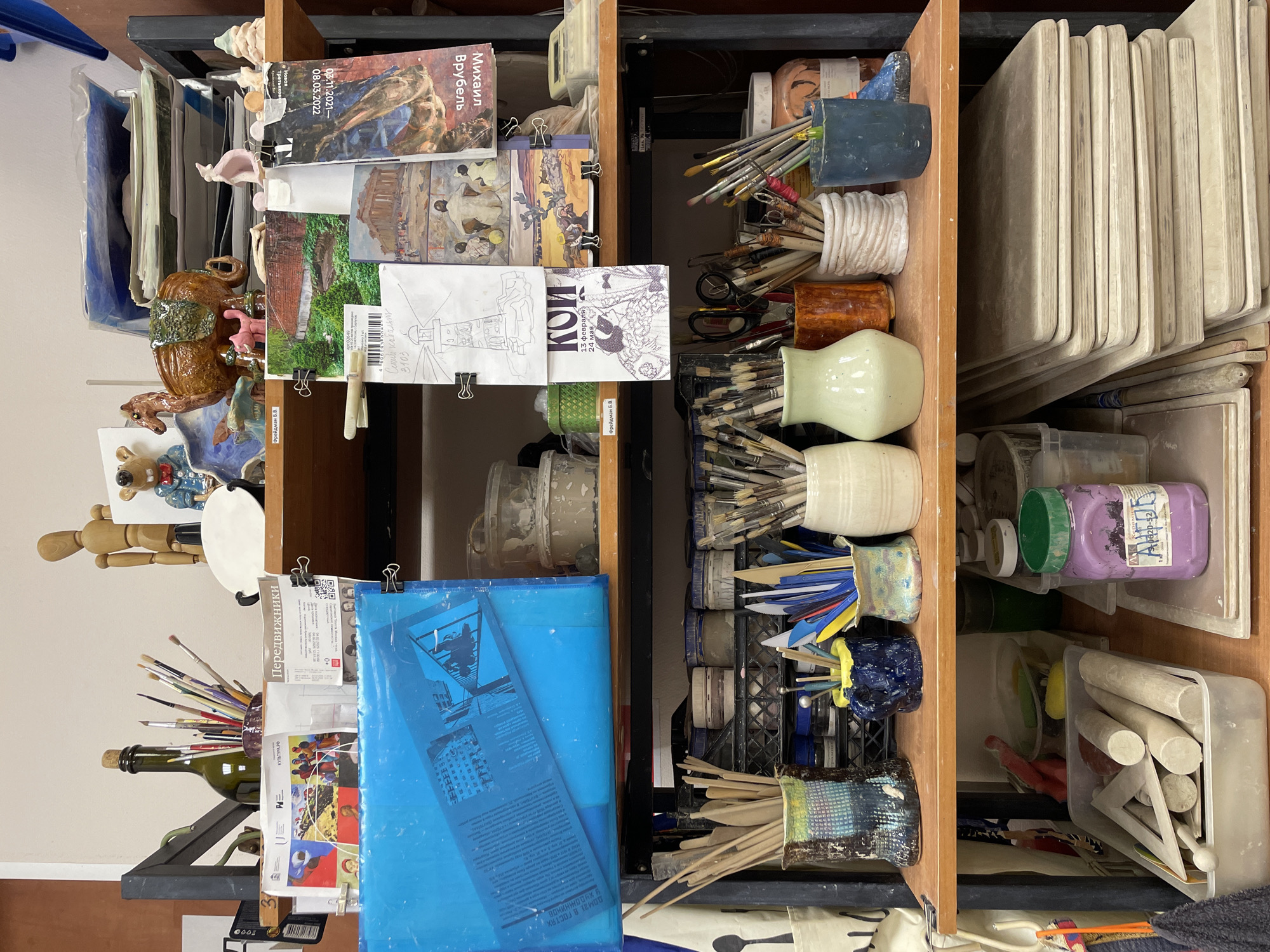 This image shows a cluttered shelf filled with art supplies. The shelf is divided into sections with a wooden frame. On the left side, there are several folders and papers, some held together with binder clips. A ceramic sculpture and a wooden mannequin are also present. In the middle section, there are various containers holding paintbrushes and tools. The right side of the shelf is filled with rectangular boards and containers with art materials. The text "Фрейдман Б.В." appears on multiple items, indicating a possible name or association. The labels on some of the items suggest a cultural context, with references to Mikhail Vrubel, an art exhibition called "Передвижники", and a mention of "Кои". The location is implied by the Russian text on various items, but specific information about the city of Khimki, Russia, is not directly visible in the image.