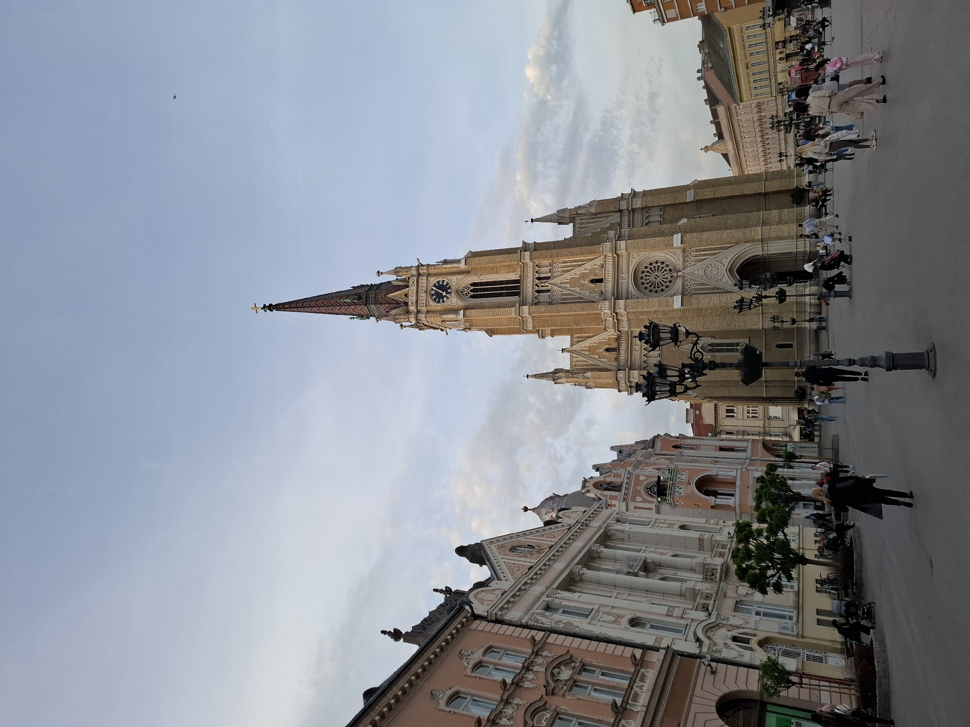 This vertical image captures a vibrant public square in Novi Sad, Serbia, featuring prominent architectural landmarks under a partly cloudy sky.
The central focus is a grand, Gothic Revival church, identified as the Name of Mary Church (Catholic Cathedral), towering majestically with its tall, slender spire. The spire is adorned with a distinctive patterned, multi-colored roof and topped with a cross. The church's façade is intricately detailed with light-colored stone, showcasing a large, visible clock face and a prominent rose window above its main entrance.
To the right and in the foreground, several ornate buildings frame the square, exhibiting classical and Baroque architectural styles. These structures feature decorative cornices, columns, arched windows, and balconies, painted in soft tones of pink, cream, and orange, which complement the lighter stone of the cathedral.
The paved square is populated by numerous people dressed in casual attire, seen walking, standing, and interacting. Ornate, old-fashioned streetlights with multiple lanterns are spaced throughout the area, alongside a few small trees. The light suggests a late afternoon or early evening, with soft, diffused illumination and some discernible shadows. A small bird is visible flying high in the clear expanse of the sky. The scene conveys a calm yet lively atmosphere typical of a historic European city center. No specific text is clearly readable on any signs or buildings.