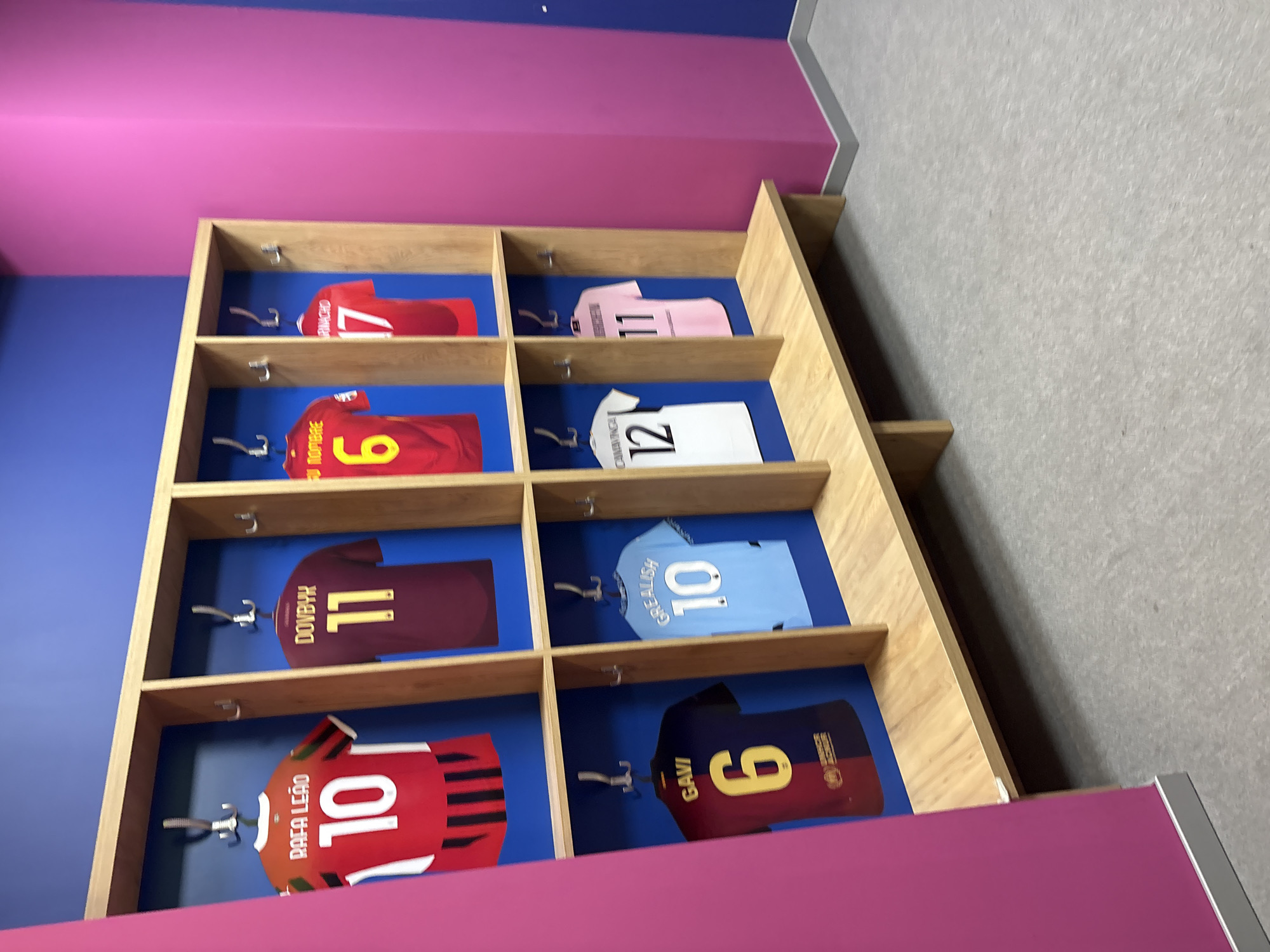 The image shows a locker room with a row of lockers. Each locker has a soccer jersey hanging inside. The jerseys are for various players, including Rafa Leão, Dovbyk, Gavi, Grealish, and others with numbers 10, 11, 6, 17, 12, and 11. The locker room has a blue wall and a pink wall. There is a wooden bench in front of the lockers. The floor is carpeted. The scene is in Valencia, Spain, at the Mestalla Stadium.