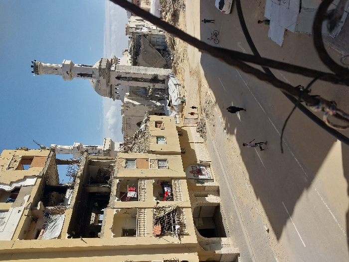 This image depicts a scene of extensive urban damage in Khan Yunis, Palestinian Territory, viewed from an elevated position. The foreground shows a dark, ornate metal railing, likely part of a balcony.
Dominating the left side is a multi-story building, light tan in color, exhibiting severe structural damage. Multiple floors are partially collapsed, with exposed interiors, shattered windows, and debris. Despite the destruction, laundry hangs on lines on some of the remaining balconies on the lower levels. Graffiti is visible on a wall section near the ground floor.
In the midground, a paved road with white lane markings stretches horizontally. Several individuals are present: A young male in a white shirt and dark pants walks on the left side of the road. Another person in dark attire walks in the center. On the right side, a person stands next to a bicycle, near a white four-door sedan. Further right, a white tent structure is partially visible, with a small figure nearby. In the distance, beyond the road and an area of rubble, three small figures (appearing to be children) walk on a dirt path.
The background is dominated by a large, white mosque with a prominent dome and two minarets. The right minaret appears largely intact, while the left minaret shows significant damage or partial collapse. The area surrounding the mosque and extending into the distance is covered in extensive rubble and debris, indicating widespread destruction. The sky is clear blue with scattered white clouds. The scene is illuminated by strong sunlight, casting distinct shadows on the road.