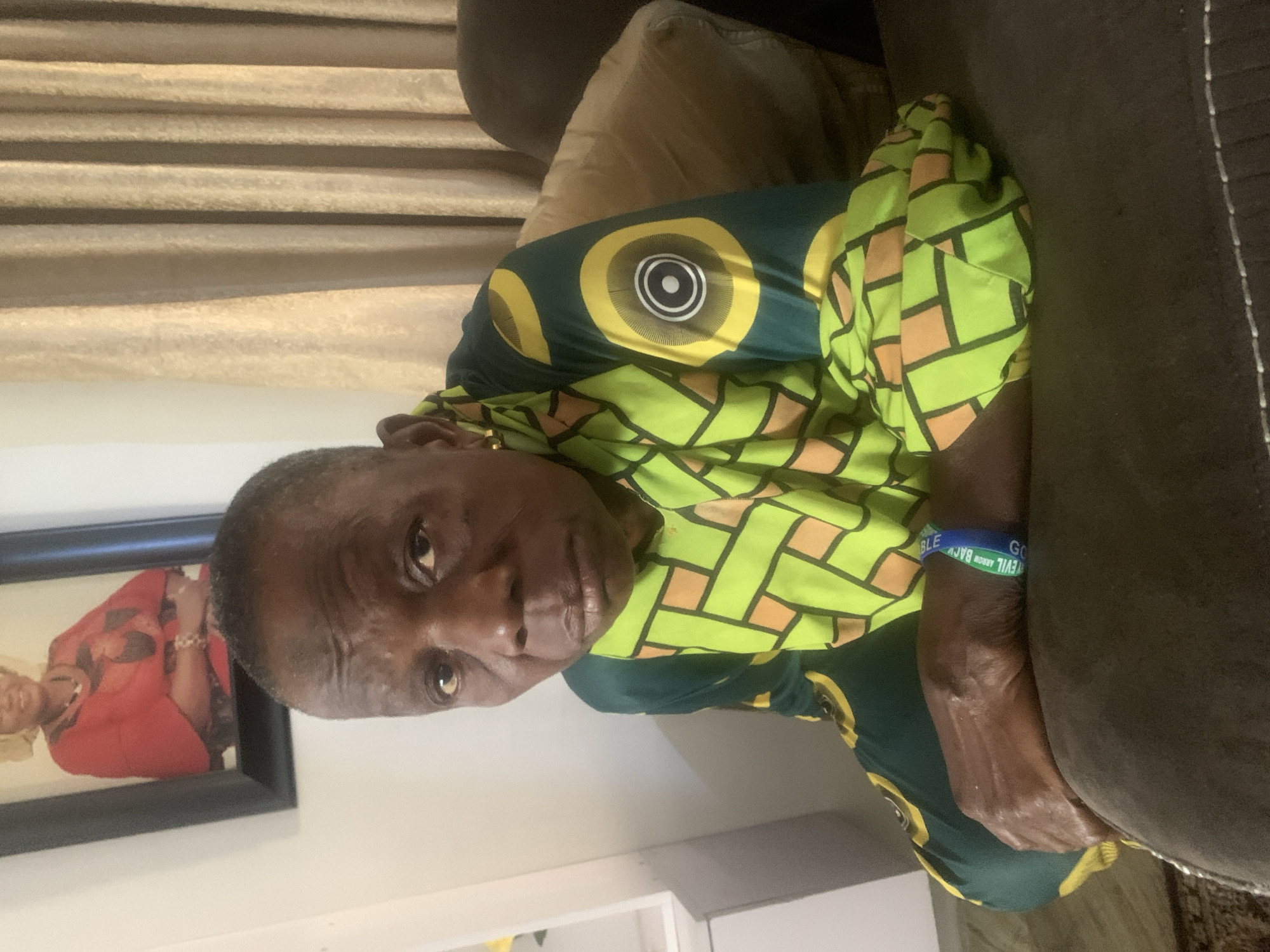 An older person, possibly female, is seated on a brown sofa in what appears to be an indoor setting, likely a living room, in Jos, Nigeria. The individual is looking directly towards the camera with a neutral expression. They are wearing a vibrant, short-sleeved shirt with a distinctive African print pattern, featuring a combination of dark green, lime green, yellow, and orange-brown colors. The pattern includes large yellow circles with concentric rings on the chest area and geometric, rectangular shapes throughout the rest of the fabric.
On the person's left wrist, a blue and green wristband is visible. Text on the wristband reads "EVIL ARROW BACK" or similar, with "EVIL" and "ARROW" clearly discernible. A gold-colored earring is visible in their left ear.
In the background, to the left of the person, a framed picture hangs on a light-colored wall, depicting another individual in red attire. Behind the sofa, light-colored curtains with horizontal pleats are visible. The lighting suggests an indoor environment, either naturally lit during the day or illuminated by artificial light sources. The overall scene conveys a calm, domestic atmosphere.