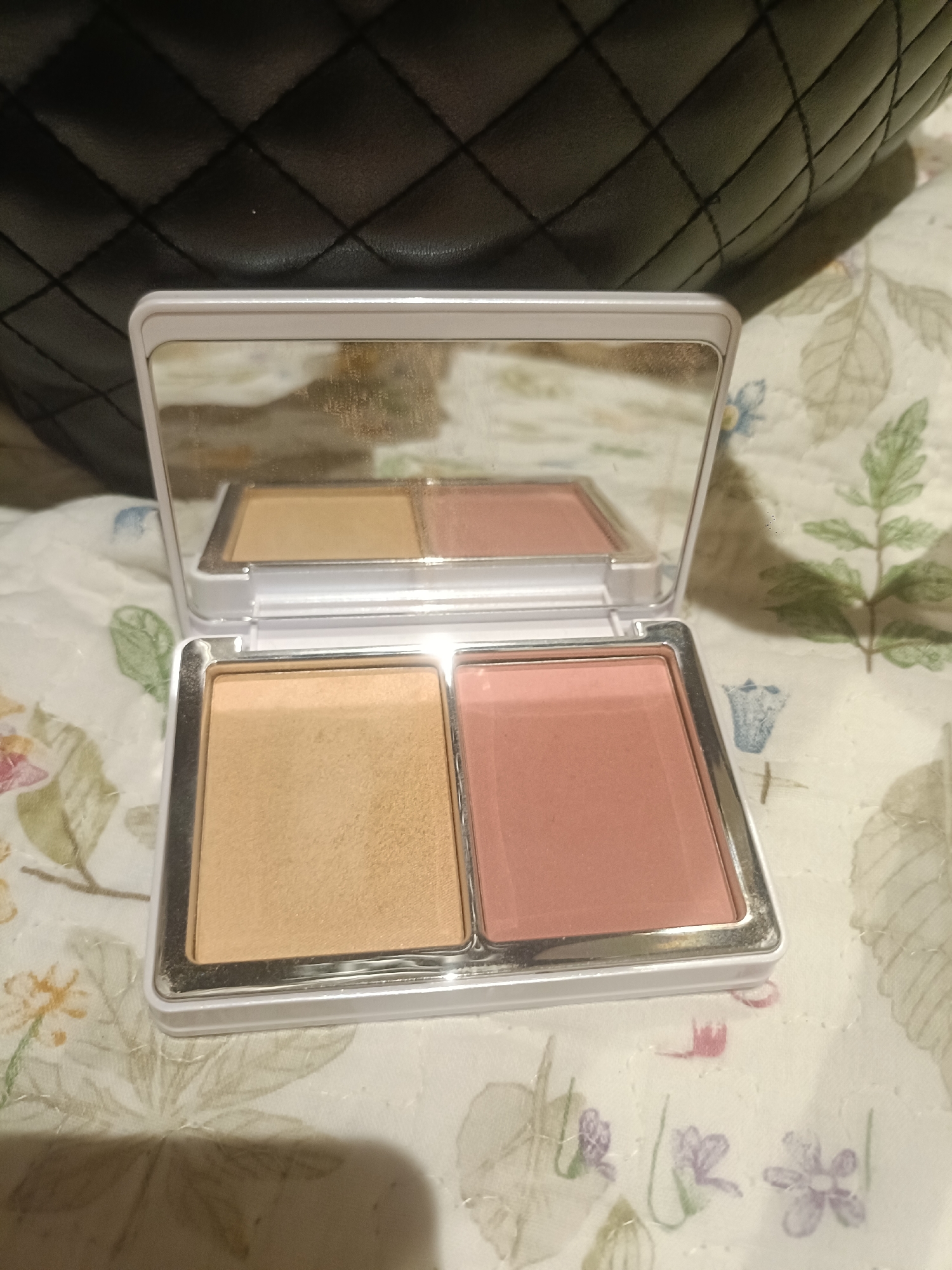 An overhead, close-up shot shows a double-paned makeup compact resting on a patterned fabric surface. The compact is open, revealing a mirrored lid and two distinct makeup shades: a light tan powder on the left and a peachy-pink blush on the right. The fabric surface is a light cream color with a repeating pattern of stylized green leaves and small purple and pink flowers. In the upper portion of the image, a black, quilted material with a diamond pattern is visible, casting a shadow over the makeup compact. The lighting appears to be artificial, with a slight sheen reflecting off the compact and its mirror. There is no indication of a specific location in Dubai or the United Arab Emirates.