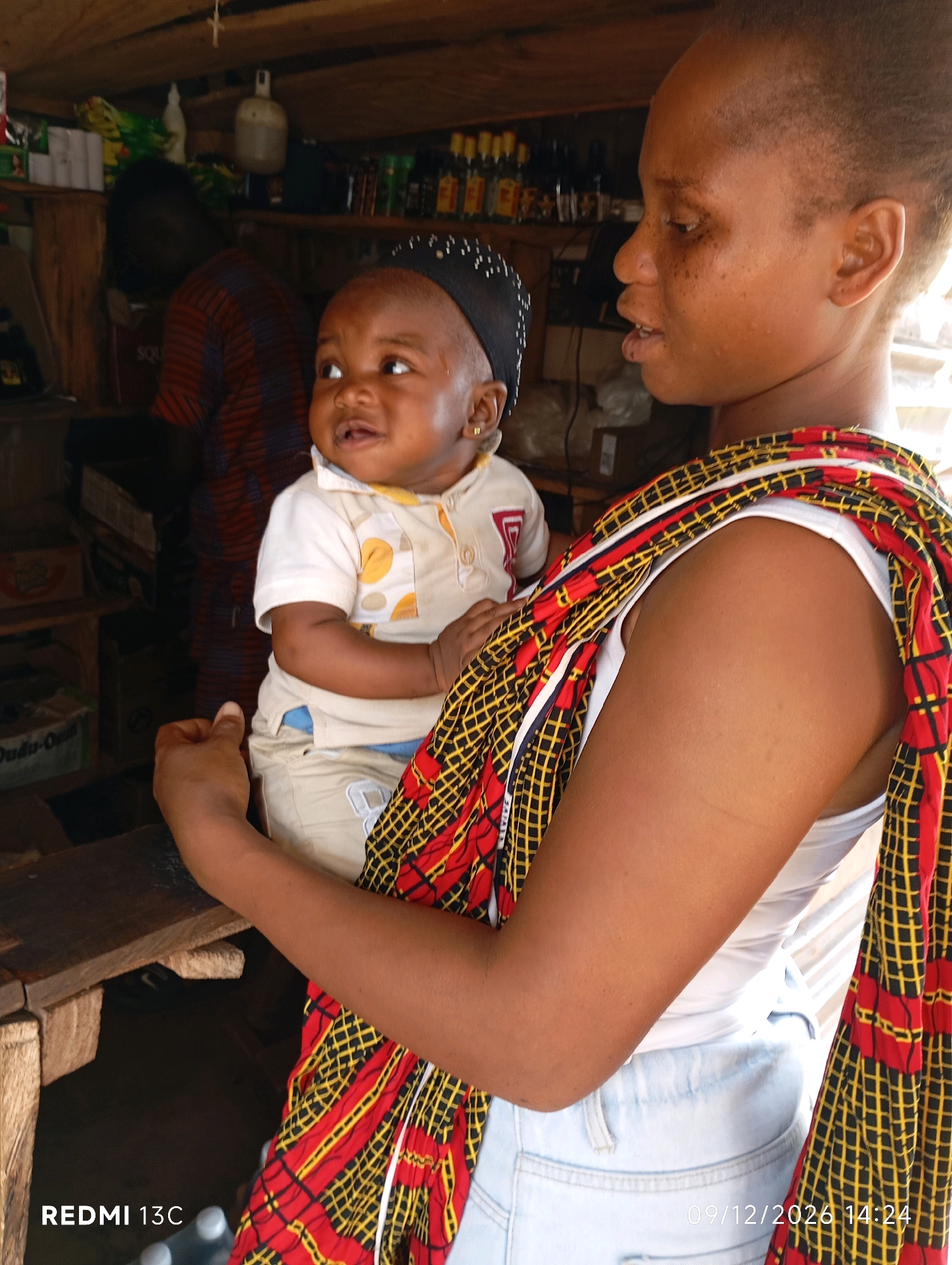 In a dimly lit interior, likely a market stall or shop in Shendam, Nigeria, a young woman cradles a baby. The woman, with dark skin and a close-cropped haircut, wears a white tank top and light blue jeans. A vibrant, patterned fabric, predominantly red and yellow with black checkered lines, is wrapped around her and the baby, serving as a sling. The baby, wearing a white polo shirt with yellow circular patterns and a black skullcap, looks upward and to the left with an open mouth. The background reveals shelves stocked with various items, including bottles and what appear to be packaged goods, suggesting a retail environment. A partially visible person in a red and brown striped shirt stands further in the background. The overall scene is rustic, with wooden structures forming the shelves and a workbench in the foreground. The lower right corner displays a timestamp: "09/12/2026 14:24" and "REDMI 13C".