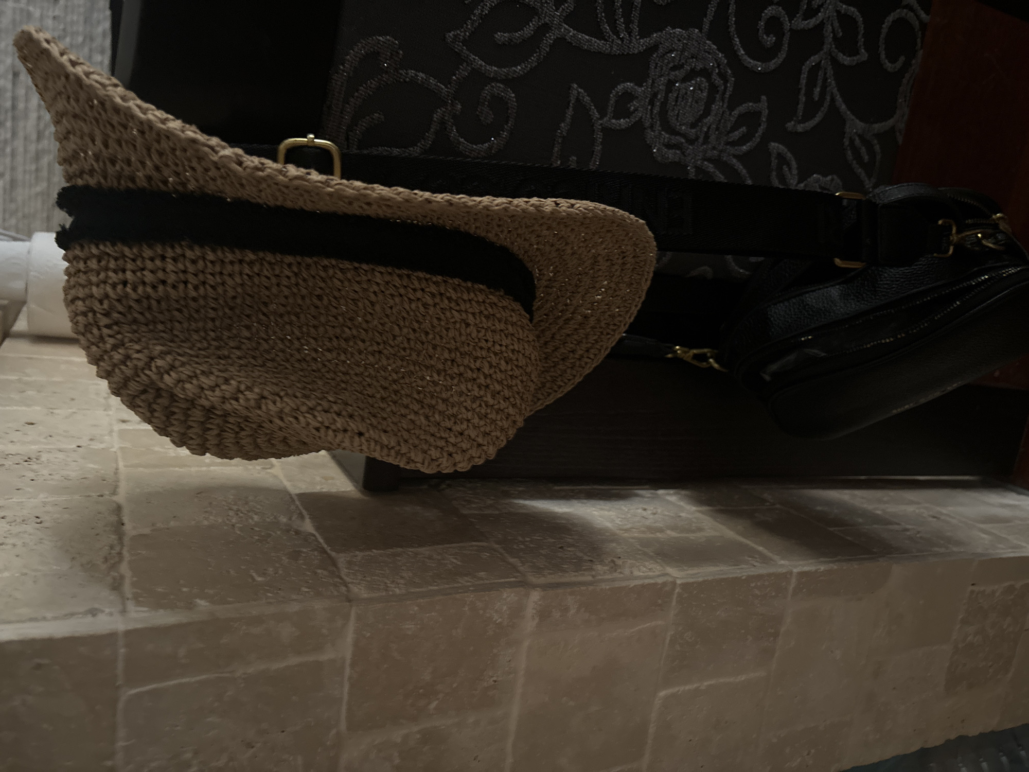 The image presents a close-up view of several items resting on a surface covered with rectangular tiles of light grey or tan color. The primary focus is a straw hat with a black band near its brim. To the right and behind the hat, there is a black bag or purse with a visible strap and metallic accents. The bag appears to be partially tucked under a dark box-like object with a floral pattern. The composition is close-cropped, and the background is out of focus, with hints of white objects partially visible in the distance to the left. The lighting in the scene seems to be muted, and there is nothing in the image that would identify the location as Budva, Montenegro.