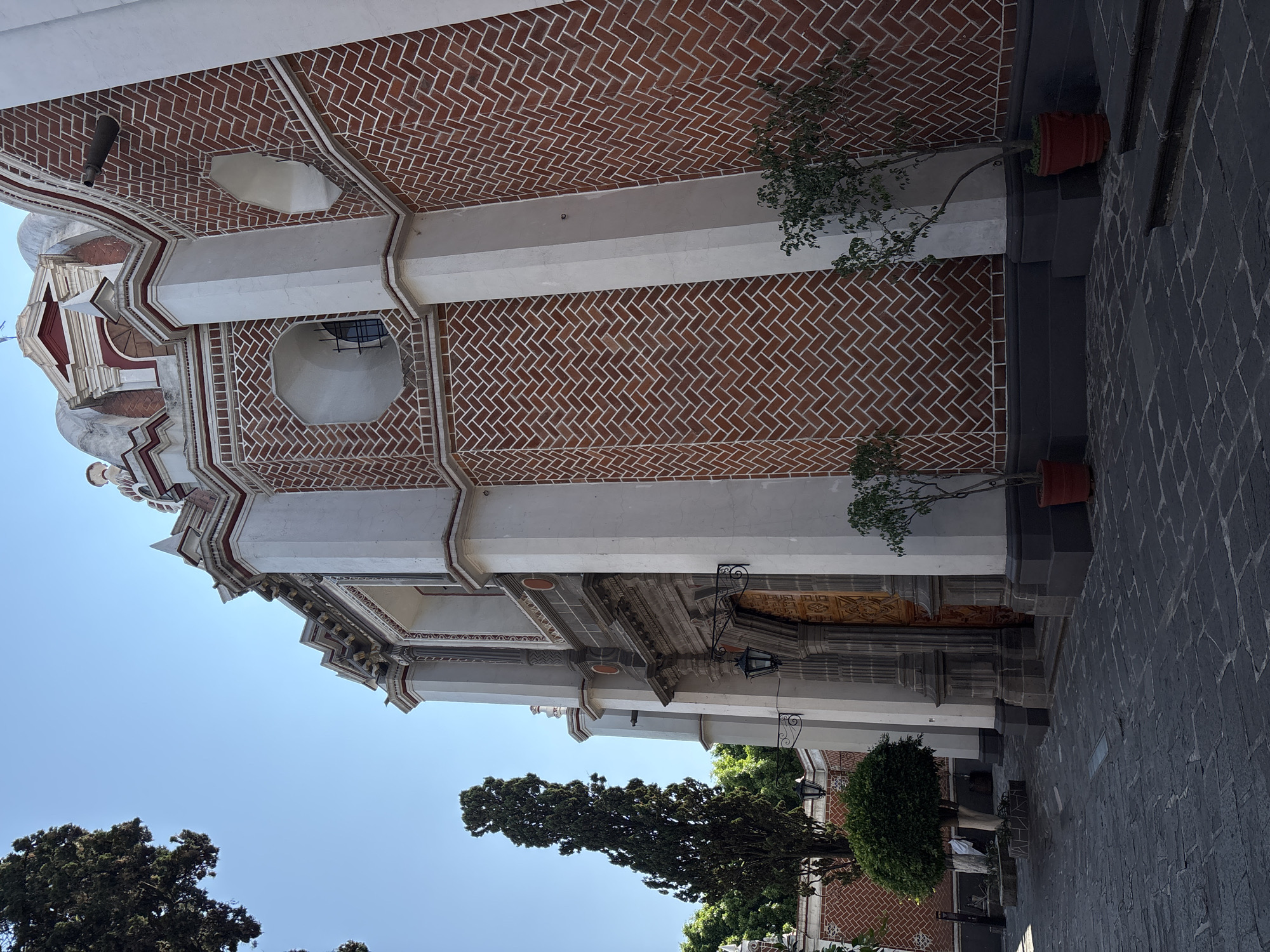 The image depicts a building in Puebla, Mexico. The architecture features a red brick facade with a herringbone pattern, interspersed with white columns and decorative elements. The building appears to be a church or similar religious structure, given its architectural style and ornamentation. Potted plants are situated along the building's exterior. The environment includes trees and a stone-paved walkway. The sky is a clear blue. There are no people visible in the image.