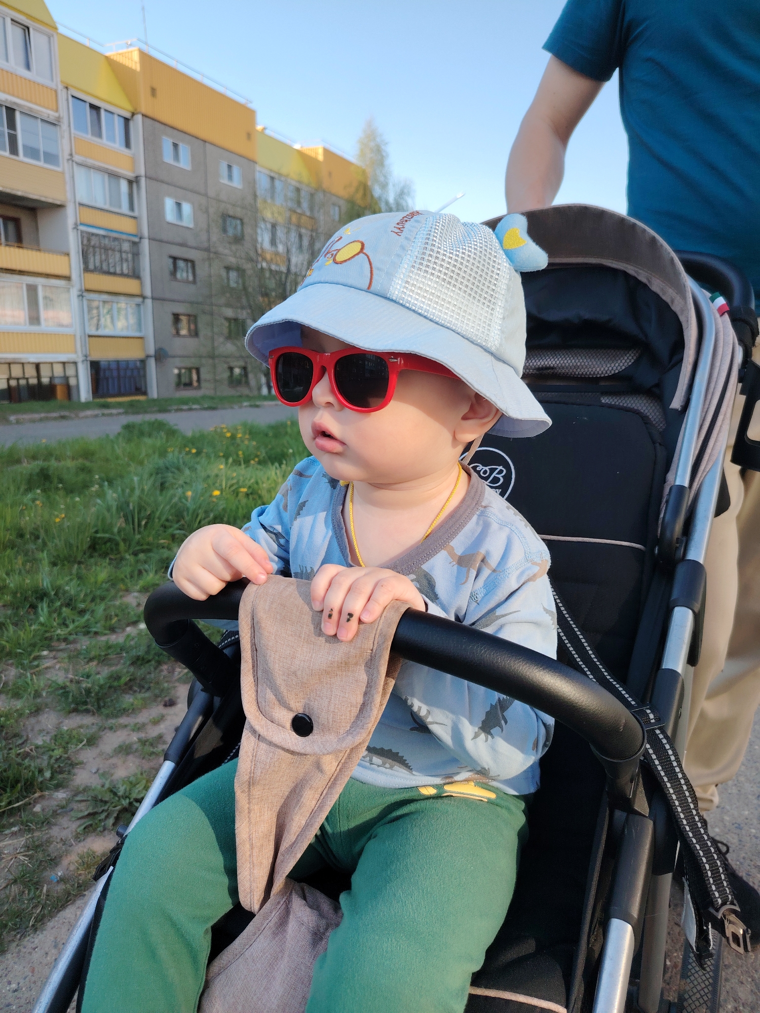 A toddler sits in a stroller on a sunny day in what appears to be an urban environment. The child is wearing red sunglasses, a light blue hat, a long-sleeved blue shirt with a dinosaur print, and green pants. The toddler's hands rest on a beige pouch attached to the stroller's handlebar. A person, likely a parent, is partially visible behind the stroller, wearing a teal shirt and beige pants. The stroller is black and silver. In the background is a multi-story building with yellow accents, indicative of a residential area. Based on the metadata, the location is Марковский, Russia. A patch of green grass is also visible in the background.