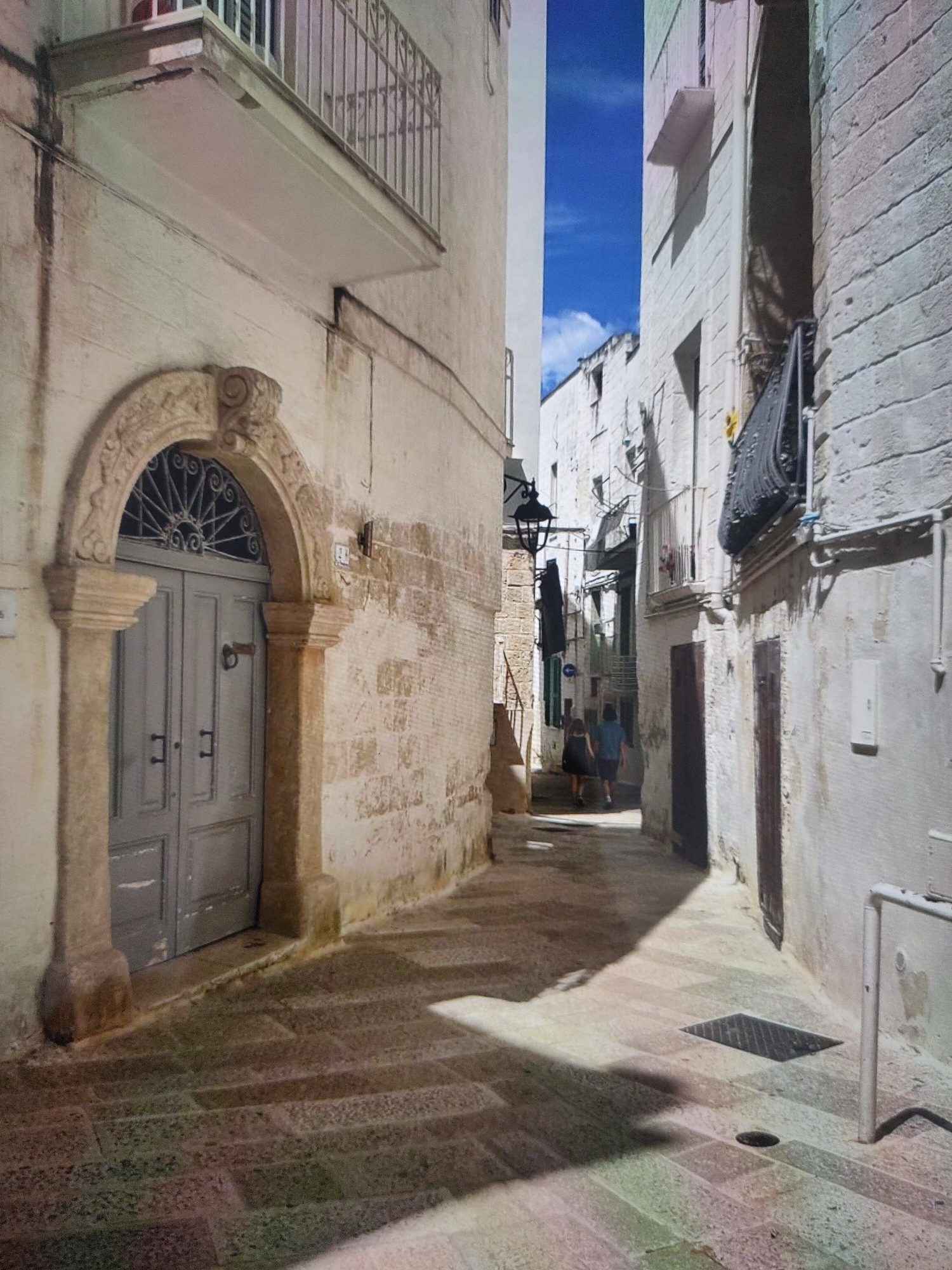 The image depicts a narrow, sun-drenched alleyway in the city of Zrenjanin, Serbia. The buildings lining the alley are constructed of light-colored stone with textured walls, suggesting an older architectural style. On the left, a prominent arched doorway with decorative stonework is visible, leading into a building. The doorway is painted a muted grey and features two dark handles and a knocker. Above the doorway, a wrought-iron grate is set into the arch. A narrow balcony with a metal railing juts out from the building on the left, casting a shadow on the wall below.
On the right side of the alley, another building displays similar stone construction and features a dark wrought-iron balcony. A black outdoor lantern is mounted high on the wall. In the distance, two figures, a woman and a man, are walking away from the camera, further down the alley. The ground is paved with large, flat stone slabs, and shadows are cast across the alley by the buildings and the sun. The sky above is a vibrant blue with a few wispy clouds. A metal grate is set into the pavement in the foreground.