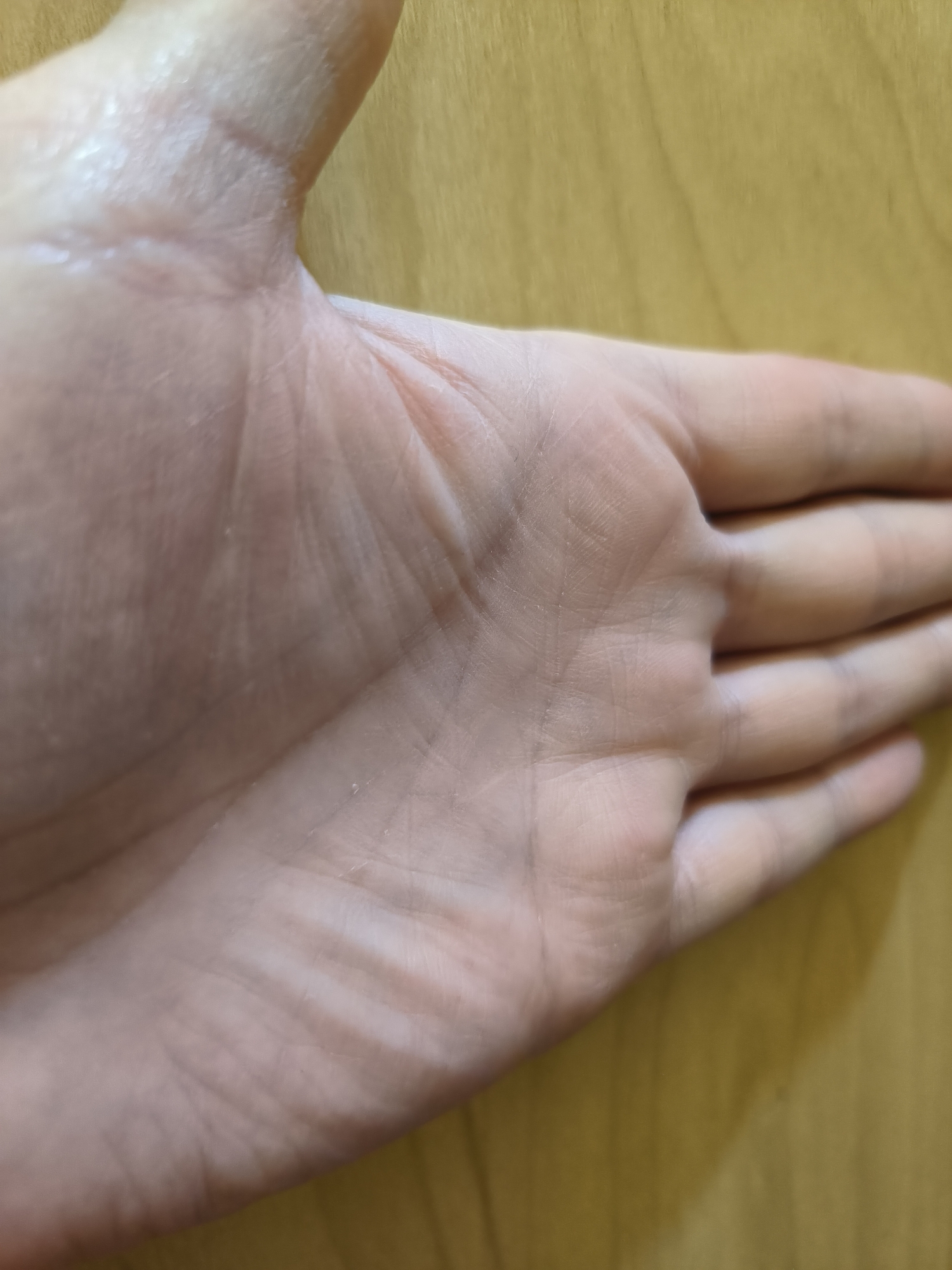 The image shows a close-up view of a human hand, palm up, resting on a wooden surface. The hand is pale with visible creases and lines on the palm, suggesting an adult. The lighting creates subtle shadows and highlights on the skin. The background is a smooth, light brown wooden surface with a faint grain pattern. The scene is static and appears to be an indoor setting. There is no information provided in the image that would allow for the identification of the location in Yuzhno-Sakhalinsk, Russia.