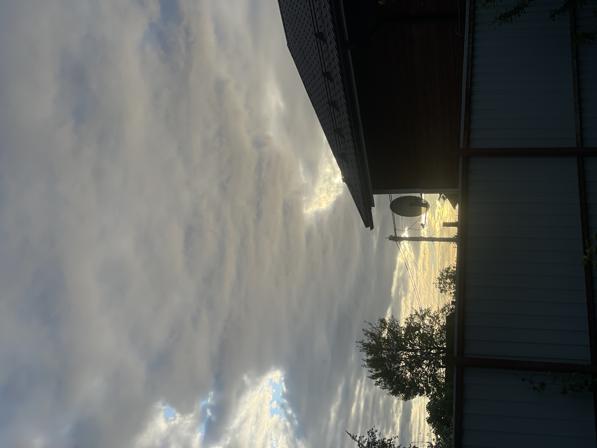 The image shows a view upwards, framed by elements of a property. A dark roof line runs diagonally from the upper left, while a light-colored metal fence with darker framing is on the right. Between these structures, there is a glimpse of a sky filled with grey and white clouds, partially obscuring the sun. Silhouetted trees and a utility pole are visible against the bright sky. There is no specific visual evidence indicating this image was taken in Sestroretsk, Russia.