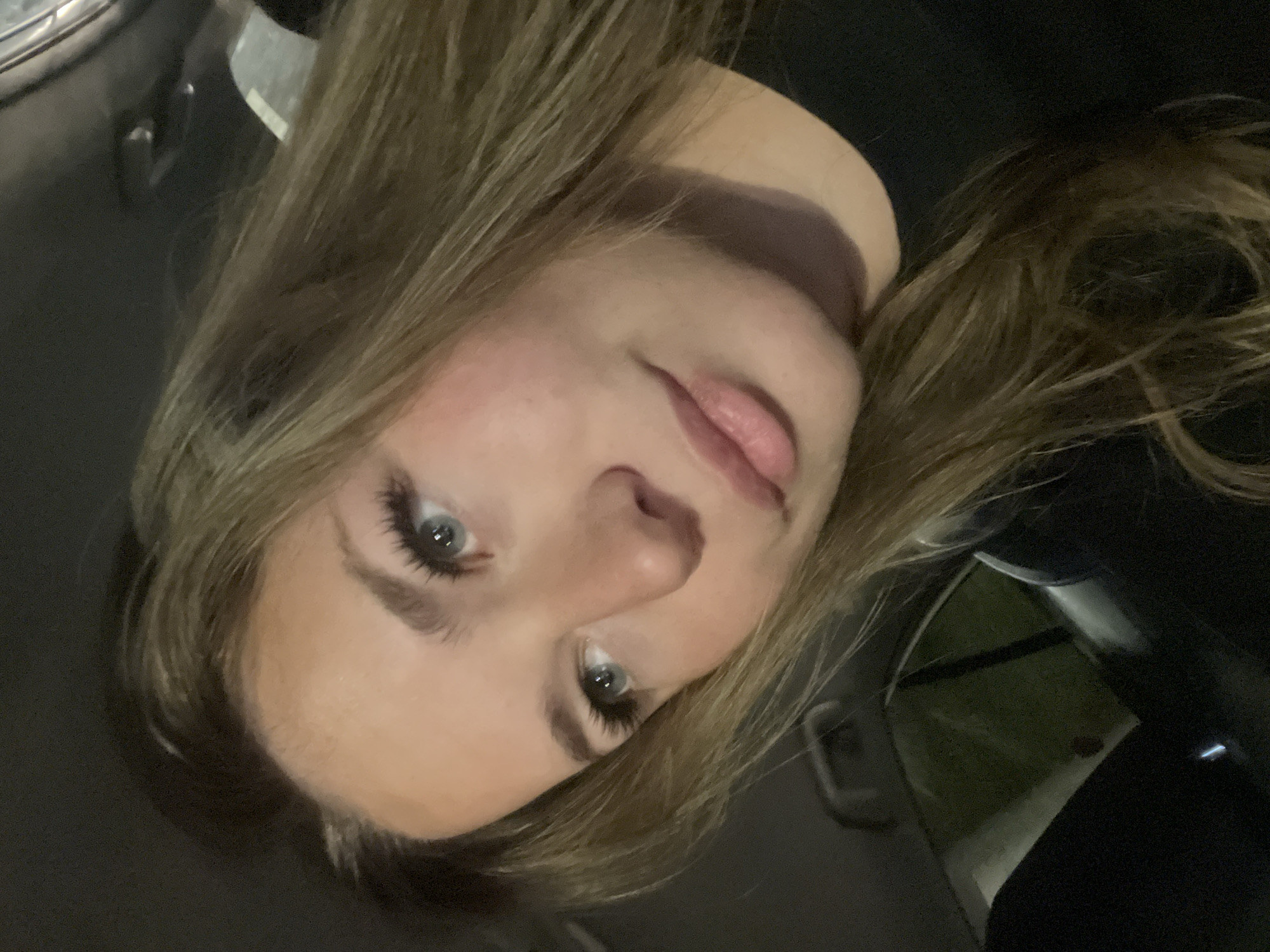 A close-up selfie of a young woman with blonde hair and blue eyes. The woman is looking directly at the camera. She appears to be inside a vehicle, as a car door and interior details are visible in the background. There is no evidence to suggest that this image was taken in the city of Budva, Montenegro.