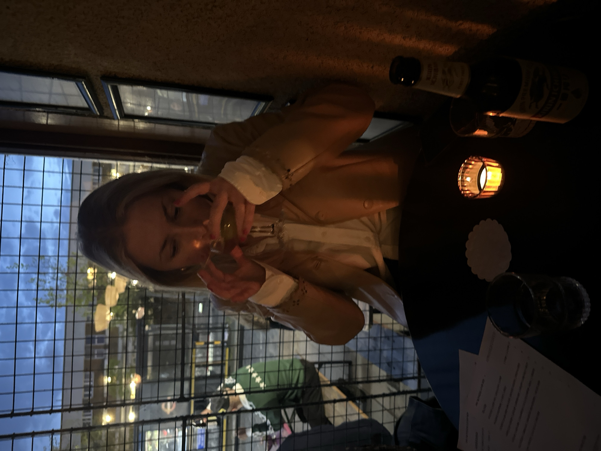 This image captures a dimly lit evening scene, likely at a bar or restaurant in London, United Kingdom, featuring a woman enjoying a drink. The main subject is a blonde woman, seen from the chest up, who is holding a glass with both hands and looking down into its contents. She is dressed in a light-colored jacket, possibly beige or light brown, over a white shirt with distinctive ruffled cuffs. Her fingers show red nail polish.
The woman is seated at a dark table where several items are arranged. A dark-colored bottle lies on its side, its label partially visible, displaying "NON-ALC" and "REAL GINGER ALE." A lit candle in a decorative metallic holder casts a warm glow on the table, illuminating a white scalloped coaster, another empty glass, and a piece of paper, which appears to be a menu with unreadable text.
The background reveals an urban setting through a metal fence or grille, likely a railing for an outdoor terrace or balcony. Beyond the fence, the overcast evening sky is visible above distant buildings, which are dotted with lights, confirming the nighttime setting. Another person wearing a green shirt is partially visible in the background behind the fence. The overall ambiance is relaxed and social, characteristic of an evening out.