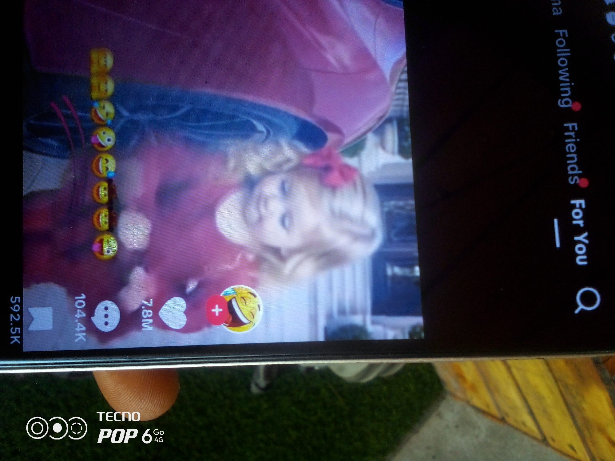 The image displays a portion of a smartphone screen, likely taken from a social media application given the presence of emojis, engagement metrics, and text elements like "Following," "Friends," and "For You." The screen is dominated by a video of a young person with blonde hair and a pink bow, appearing to be the subject of the video. Several emojis are stacked vertically on the left side of the screen, and engagement icons such as a comment bubble, a heart, and a plus sign within a laughing emoji are visible on the bottom portion. Text overlays indicate numerical engagement figures, such as "592.5K," "104.4K," and "7.8M." A finger is partially visible at the bottom of the image, suggesting the phone is being held. The background of the image beyond the phone screen appears to be a grassy surface and a wooden surface. There is no information within the image that can place the scene in Howayi, Nigeria.