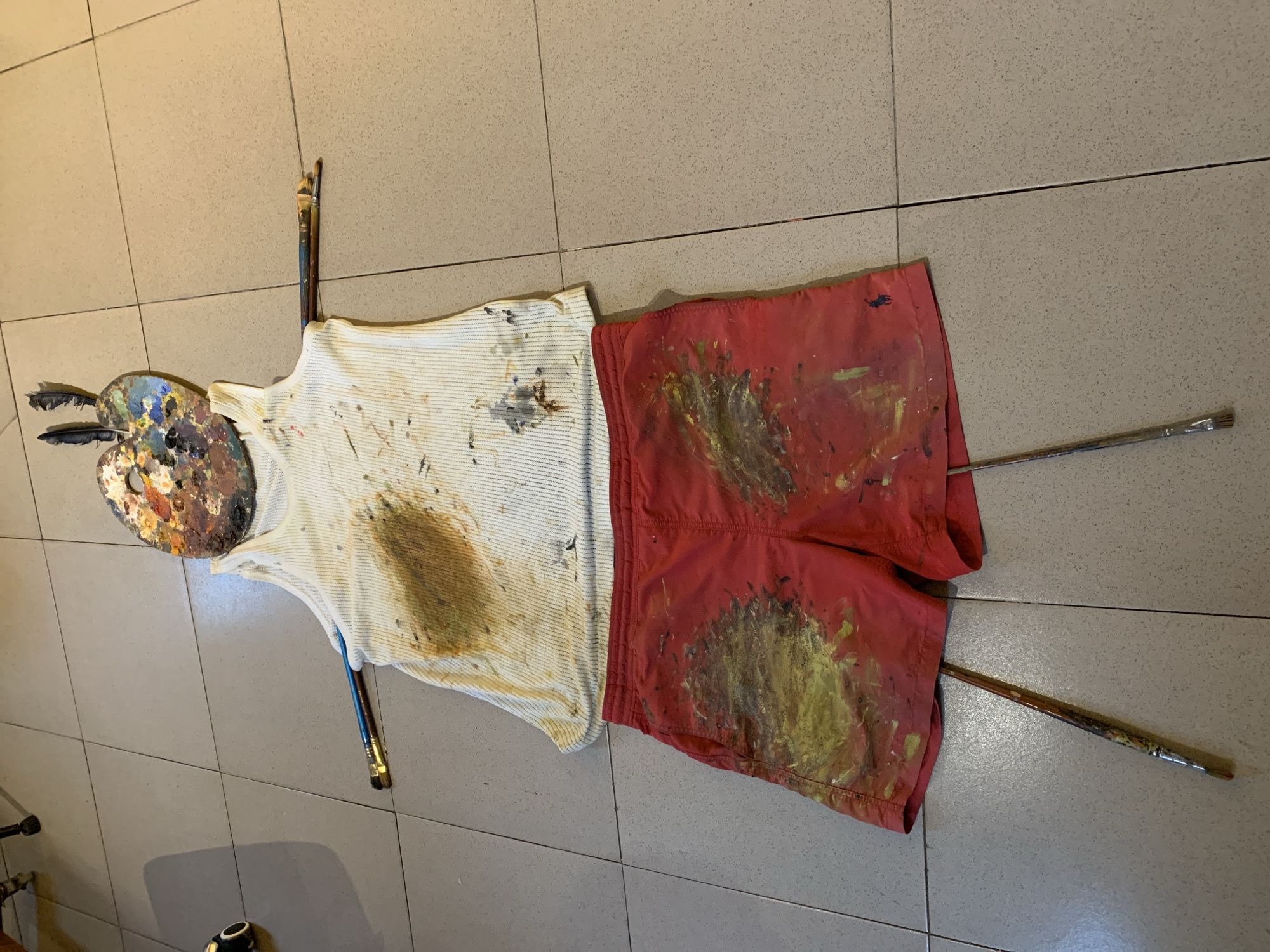 An artist's discarded outfit, a white ribbed tank top and red athletic shorts, is laid out on a beige tiled floor, appearing to be in a studio or outdoor space in Ado, Nigeria. Both garments are heavily splattered with various colors of paint, including brown, yellow, and blue. The tank top has a large brown stain on the chest. The red shorts have prominent yellow and brown paint marks. A painter's palette, also covered in dried paint, is positioned at the top of the tank top, with two black feathers sticking out of it, resembling a head. Two paintbrushes are inserted through the armholes of the tank top, extending outwards like arms. Two more paintbrushes are positioned as legs, extending downwards from the shorts. The scene suggests a creative process has recently concluded, leaving behind the artist's tools and attire.