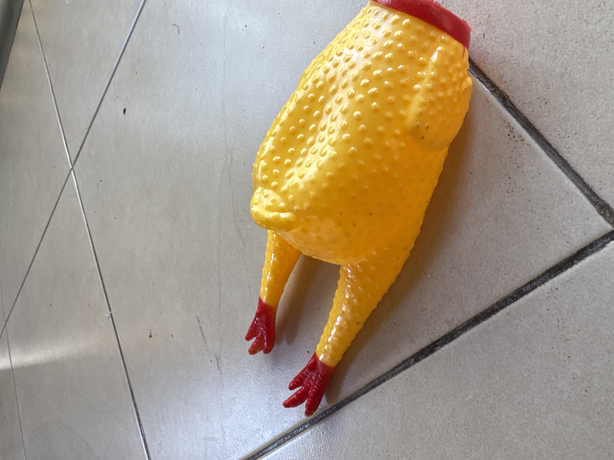 A yellow rubber chicken toy with red extremities sits on a beige tile floor with gray grout lines. The toy is situated in Kuta Selatan, Indonesia.