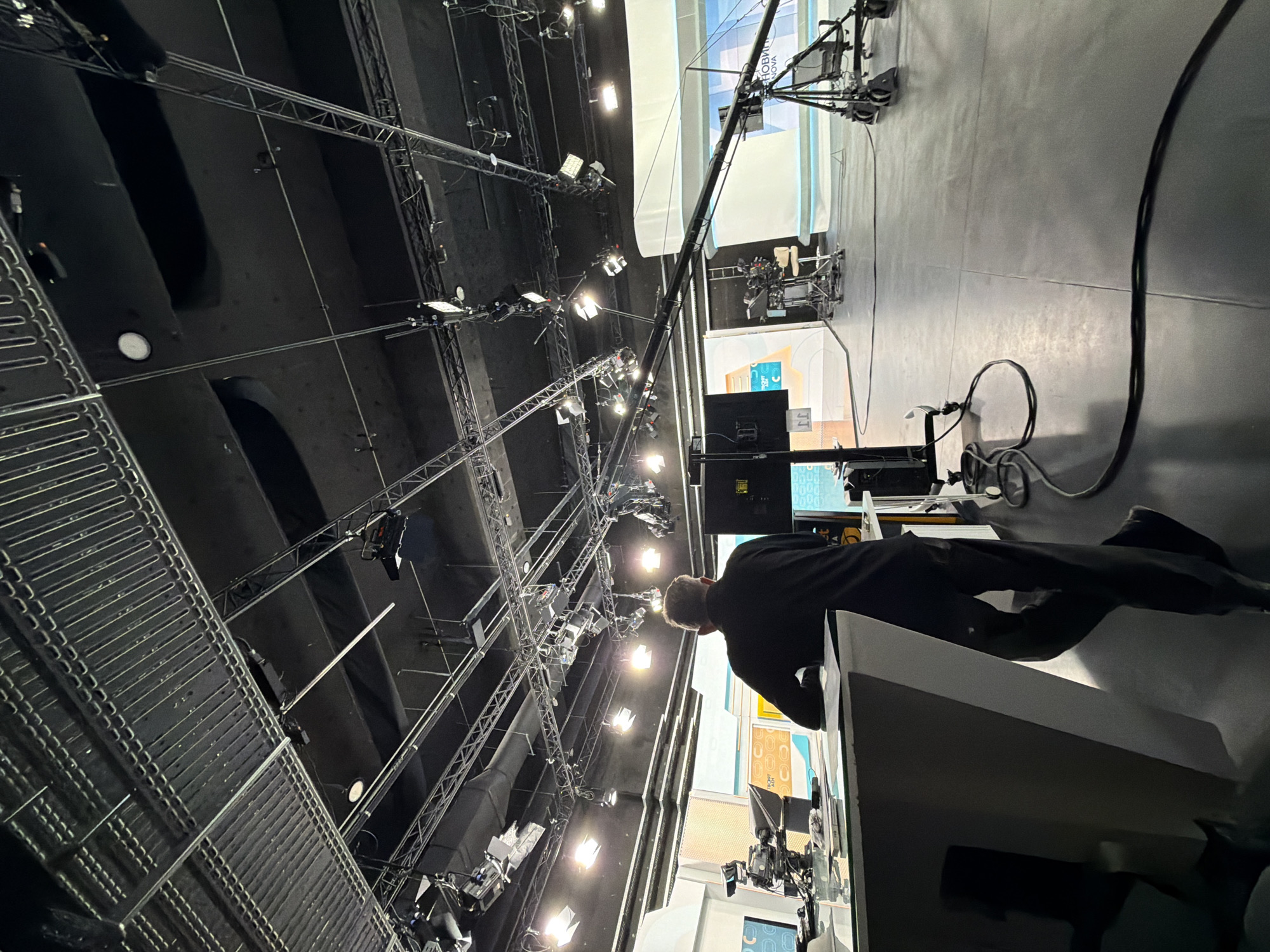 A man in dark clothing stands in a television studio environment, looking toward multiple cameras and lighting rigs. The studio has a high ceiling with complex rigging and numerous bright lights. A camera crane is visible in the background, and the floor appears to be concrete. The studio is likely located in Sofia, Bulgaria.