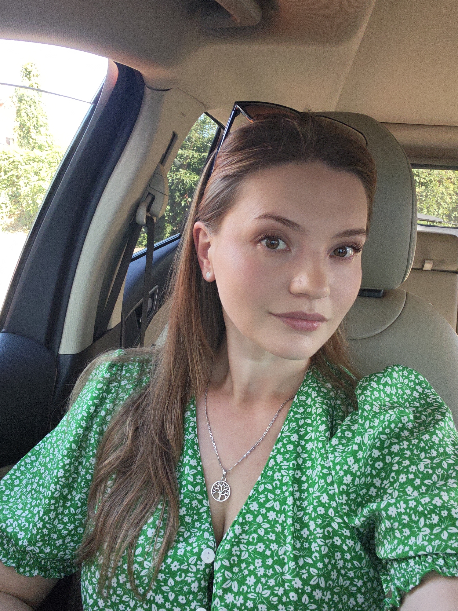 A woman is captured in a selfie taken from inside a car. She is wearing a green floral dress with puffed sleeves and a silver necklace featuring a tree of life pendant. Her hair is long and brown, and she has on a small white earring. She is looking directly at the camera with a slight smile. Sunglasses are perched on top of her head. The interior of the car is visible, including the beige leather seat and part of the car's roof lining. The background outside the car window shows lush green trees and foliage, suggesting a sunny day. The setting is consistent with residential areas or parklands within Vinnytsia, Ukraine, where the sunlight filtering through the trees indicates it is daytime.