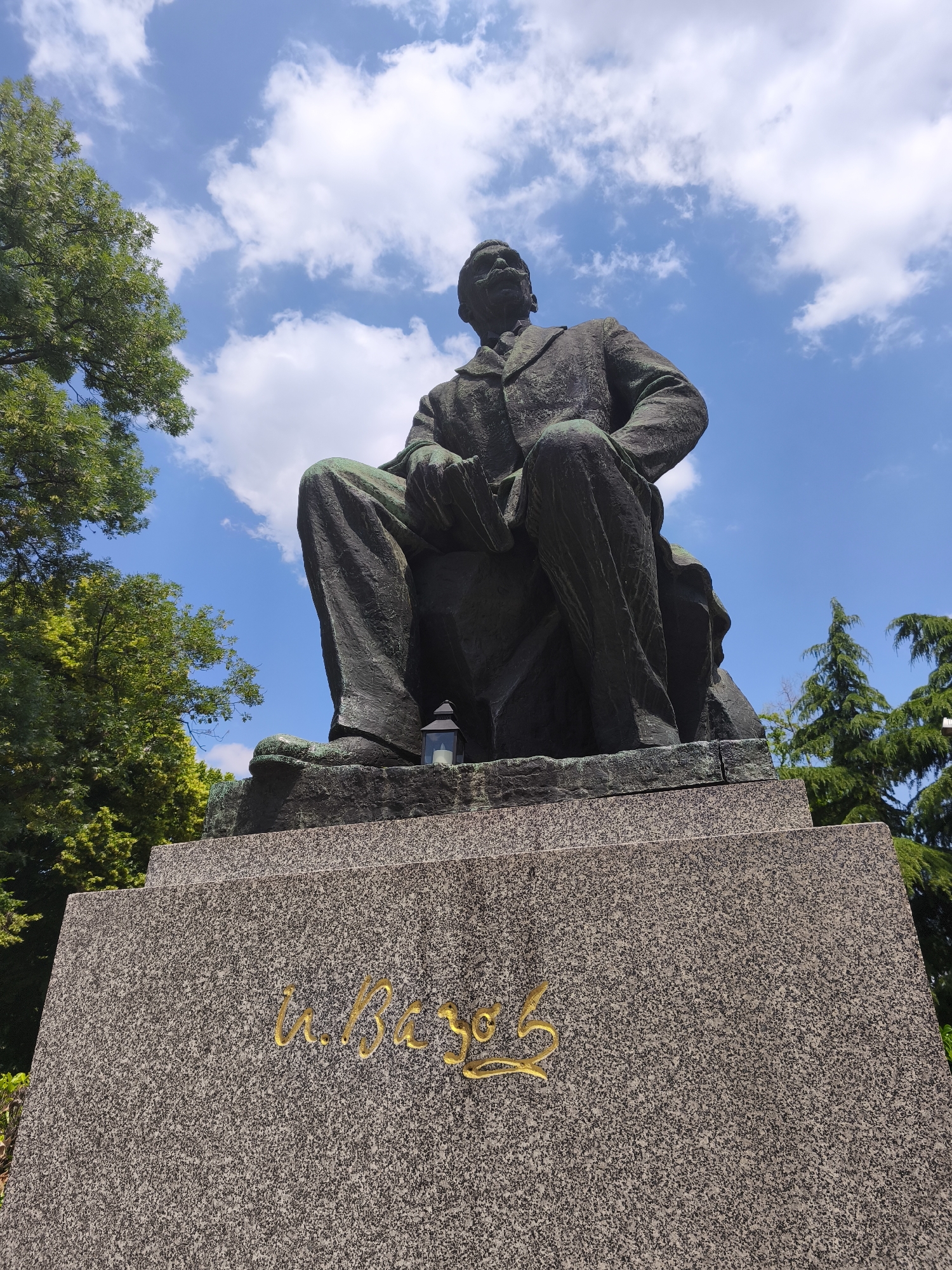 A bronze statue of Ivan Vazov, the renowned Bulgarian poet and writer, is depicted from a low-angle perspective against a bright blue sky with scattered white cumulus clouds. The statue is located in Sofia, Bulgaria, commonly found in front of the Ivan Vazov National Theatre. The monument portrays Vazov seated, his figure exhibiting a green patina, dressed in a formal suit jacket, trousers, and tie, with a prominent mustache. His head is turned slightly, and his gaze is directed upwards and to his proper right. The statue rests upon a multi-tiered pedestal constructed of speckled grey granite. On the front face of the pedestal, the name "И. Вазовъ" (I. Vazov) is inscribed in stylized gold Cyrillic script. A small, dark lantern containing a lit candle is placed on the pedestal near the statue's proper right foot. Lush green deciduous trees frame the left side of the scene, while darker green coniferous trees are visible in the background on the right. The entire scene is brightly illuminated by natural daylight.