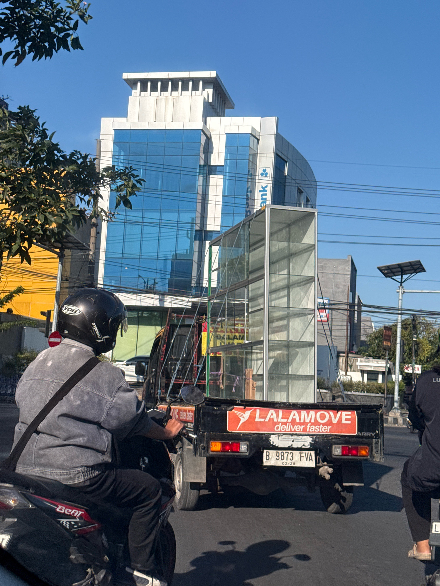 The image captures a street scene in Surabaya, Indonesia, on a clear day with a bright blue sky. In the foreground, a person wearing a black helmet and a denim jacket rides a motorcycle. Behind them, a black Lalamove delivery truck is prominently featured, carrying a large, transparent glass structure. The truck has a license plate that reads "B 9873 FVA". In the background, a modern, multi-story building with blue glass windows stands tall, with a logo that appears to be for a bank visible on its facade. The street is paved, and there are other vehicles and elements of urban infrastructure, such as utility poles and wires, visible. The lighting suggests it is daytime, with strong sunlight casting shadows.