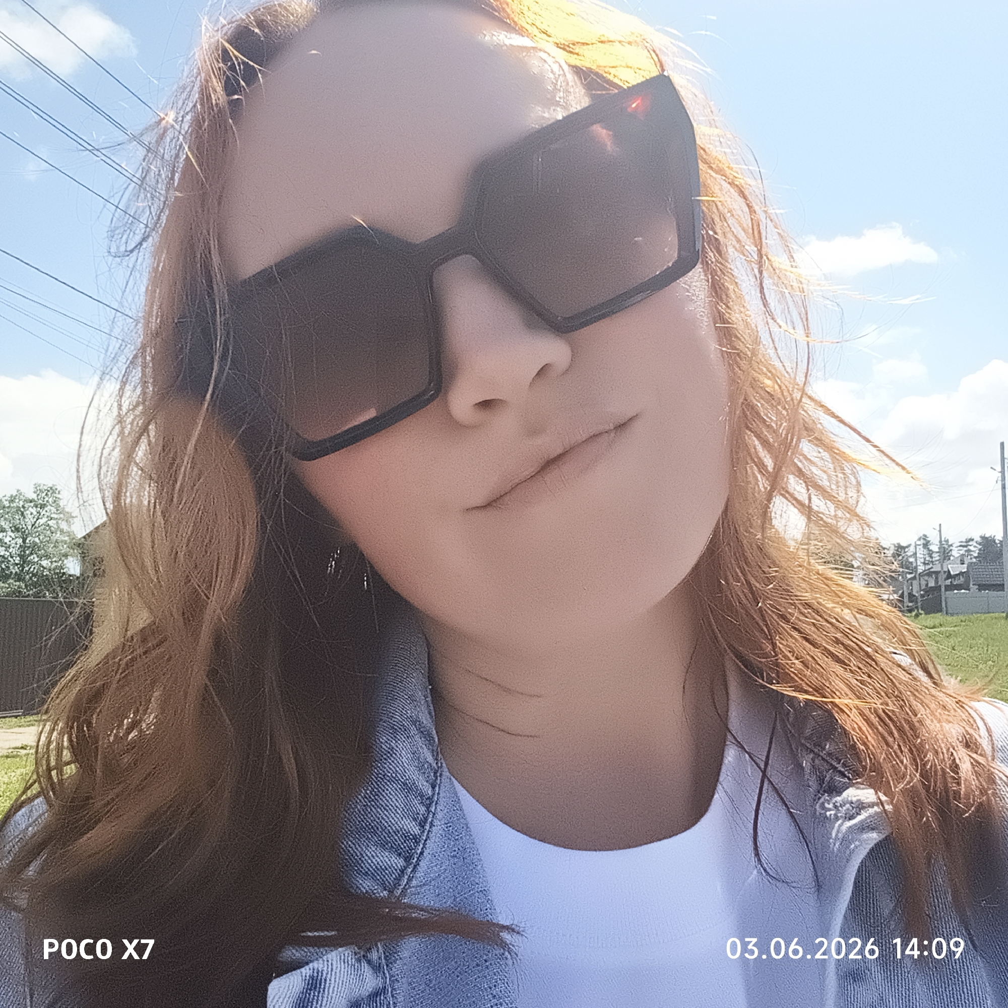 A woman is featured in a close-up selfie taken outdoors in Chaikovsky, Russia. She is shown from the chest up, looking slightly upwards and to the right, with her head tilted. Her long, reddish-brown hair appears windswept and is brightly illuminated by direct sunlight, particularly around her head and shoulders. She wears large, dark, rectangular sunglasses that obscure her eyes, and a light blue denim jacket over a white top. Her lips are slightly parted, suggesting a relaxed or pensive expression.
The background shows a clear, bright blue sky with scattered white clouds. Several power lines are visible stretching across the upper left portion of the image. In the mid-ground and distance, green grassy areas, trees, and some fencing or residential structures can be seen, indicating a suburban or semi-rural environment.
The photograph was taken on a sunny day, as evidenced by the strong lighting and the woman wearing sunglasses. A digital timestamp in the bottom right corner reads "03.06.2026 14:09," indicating the date and time of capture. The device used is identified by "POCO X7" in the bottom left corner.