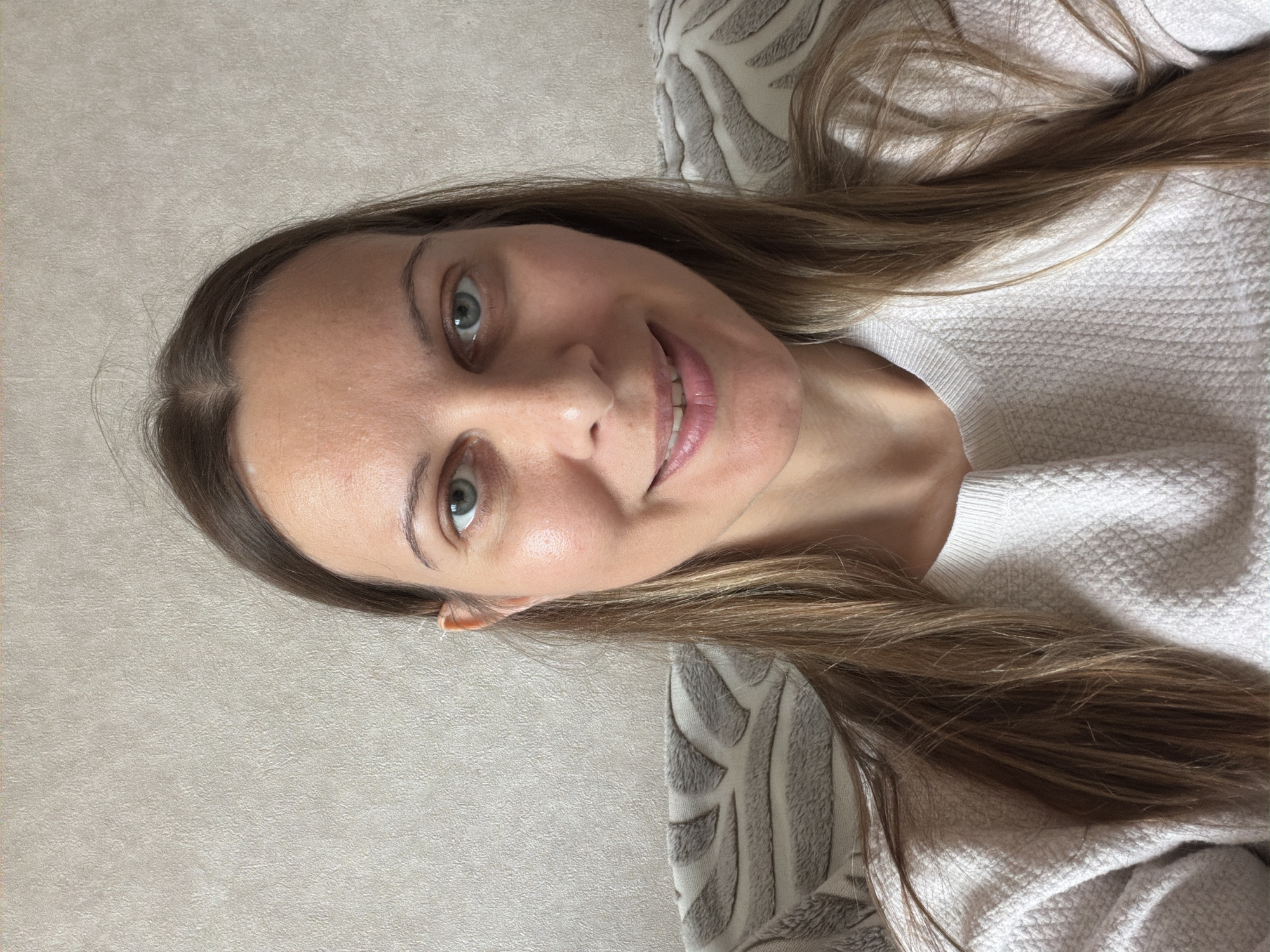 A woman with long brown hair and blue eyes smiles at the camera. She is wearing a light-colored, textured sweater. She is seated in front of a wall with a textured pattern, and part of a patterned cushion is visible on her right. There are no other individuals or objects of note in the frame. The scene appears to be indoors, likely in a residential setting. The provided context of Izhevsk, Russia, does not offer any specific visual cues to confirm or deny this location.