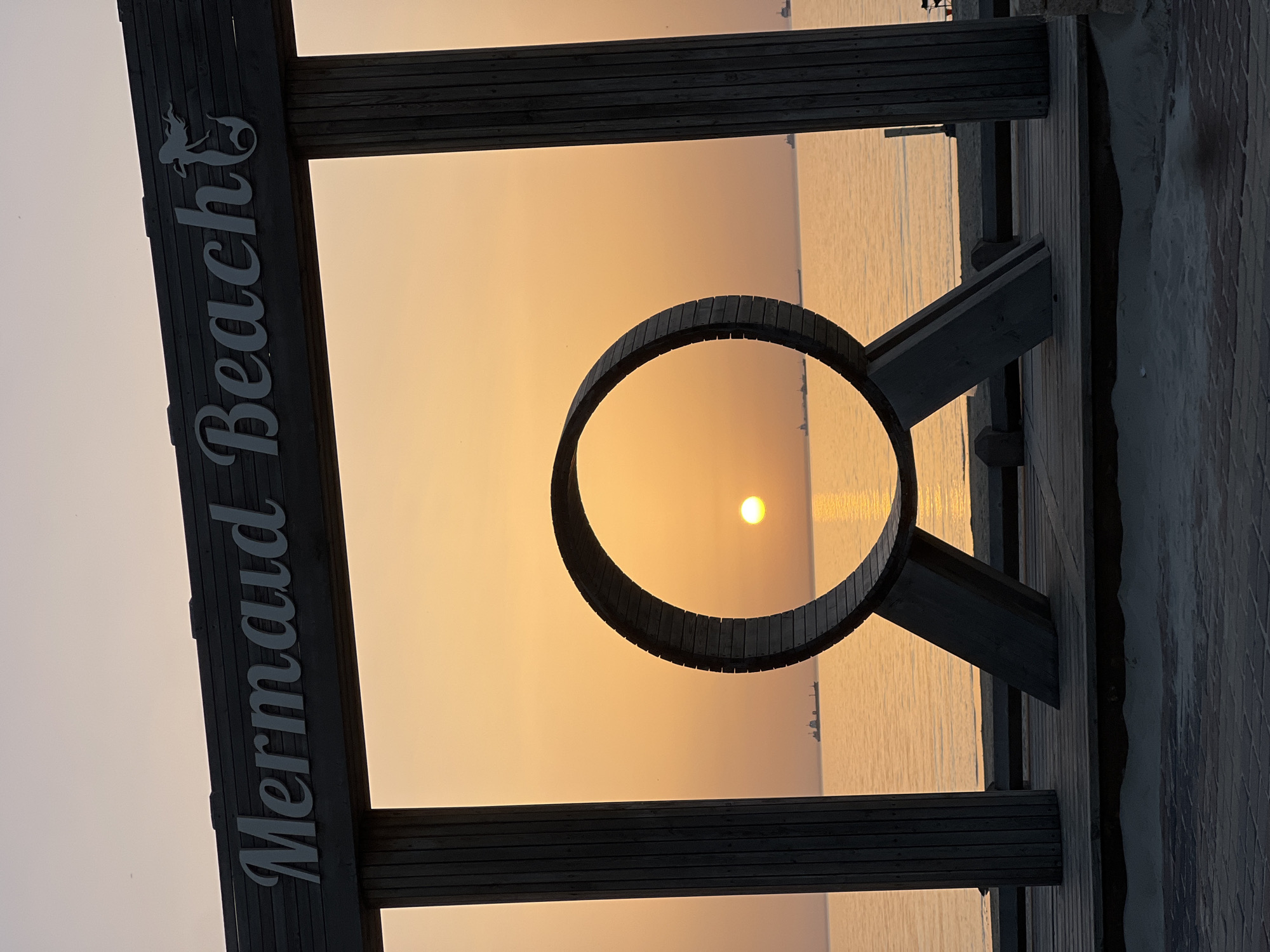 The image, taken in Kiryat Yam, Israel, depicts a wooden structure framing a sunrise or sunset over the sea. The structure includes a rectangular frame with the words "Mermaid Beach" vertically along the left side. A circular, segmented wooden ring is attached to the frame's right side, creating an additional framing element. The sun is positioned within the ring's center, casting a reflection on the water's surface. The water appears calm. In the background, distant ships are visible. The lighting indicates the early morning or late evening hours. The photograph is taken from a low angle, looking upward toward the sky and sea. A wooden boardwalk is visible on the right side of the frame.